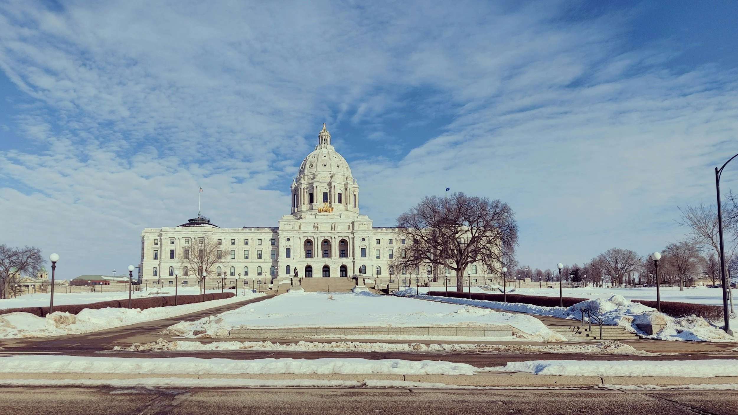 View of a large government building with a white dome, surrounded by snow-covered ground and leafless trees, under a partly cloudy sky.