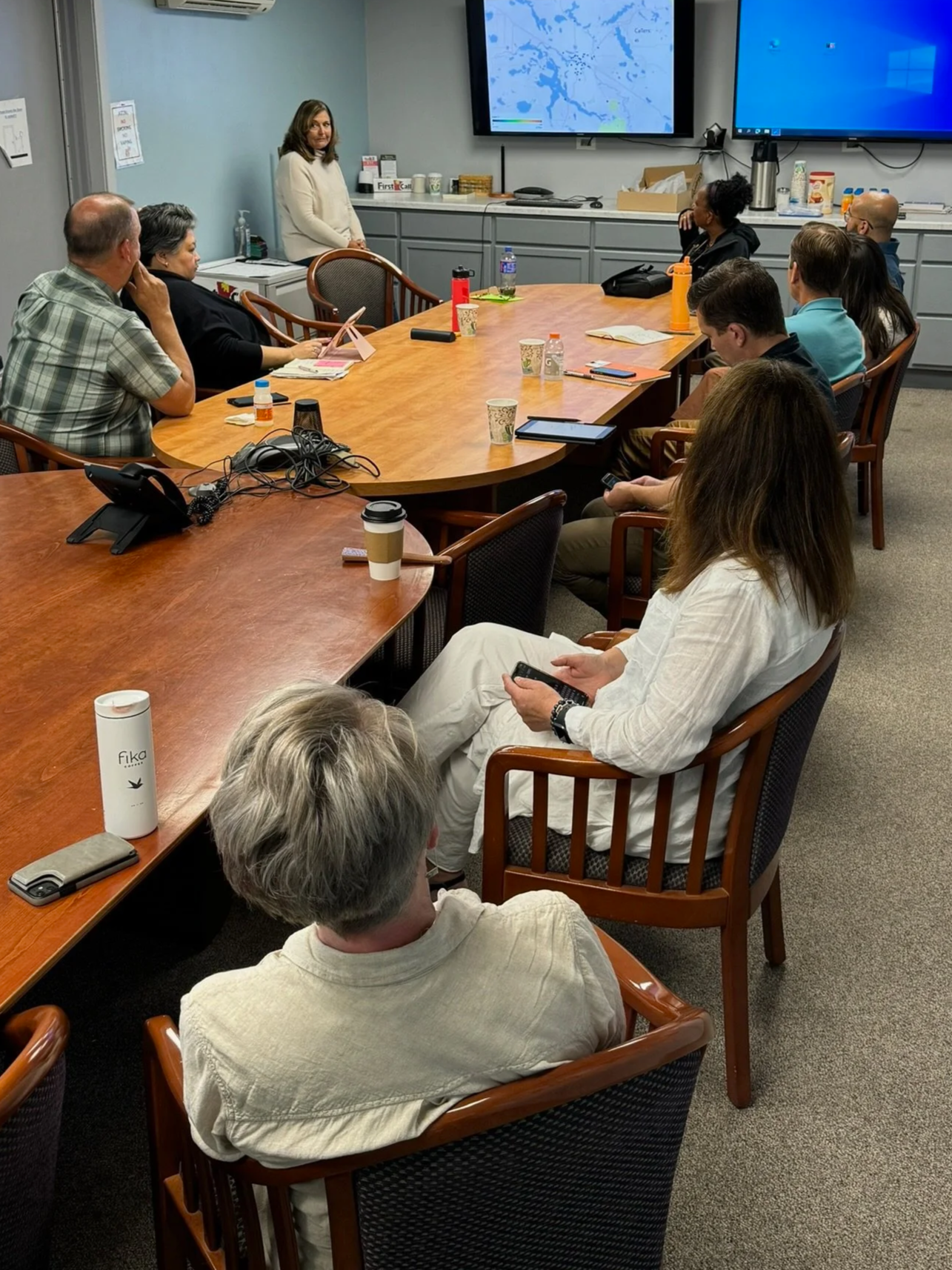 A woman is giving a presentation to a group of people seated around a conference table in a meeting room. There is a large screen behind her displaying a map, and other electronic devices and beverages are on the table.