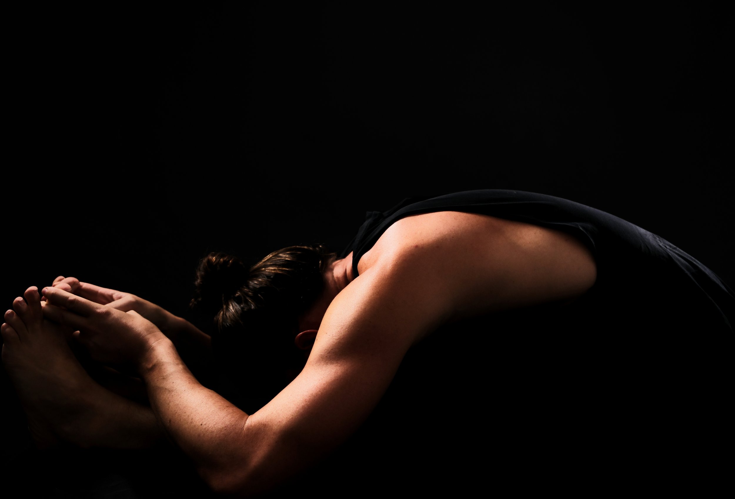 Person in a black tank top performing a forward bend yoga pose on a black background.