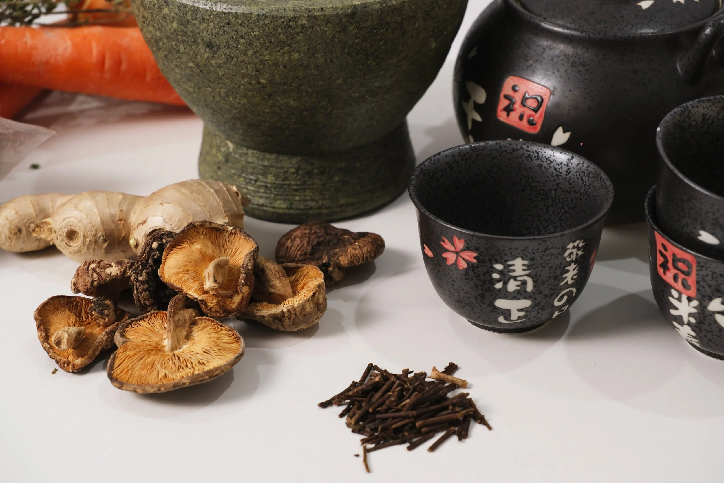 Assorted dried mushrooms, ginger root, and a small pile of dried twigs or sticks, with ceramic tea cups and bottles decorated with Japanese characters and cherry blossom designs in the background. An image of macrobiotic foods display