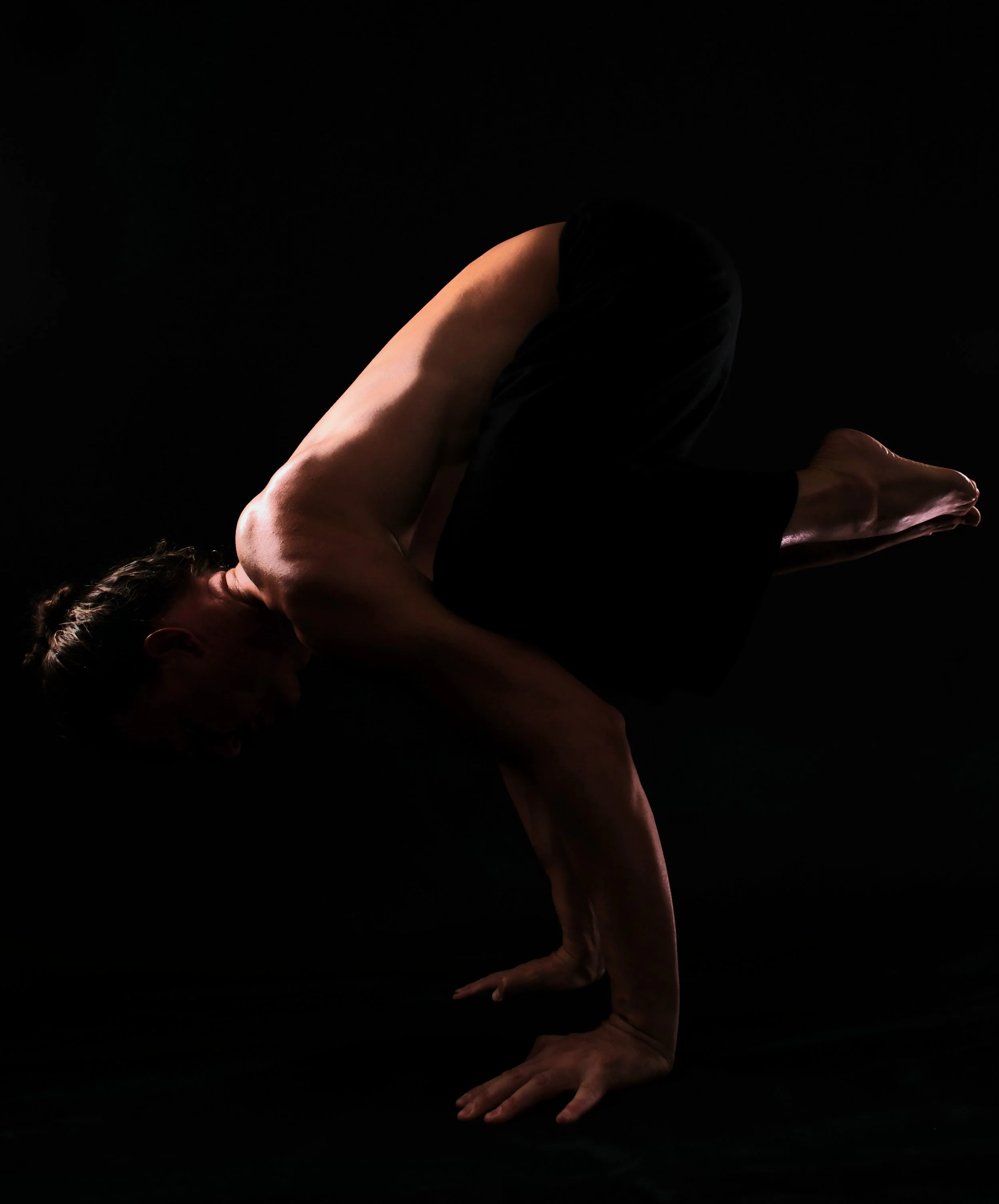 A person performing a yoga pose called the Crow Pose, balancing on their hands with knees resting on elbows, against a dark background.