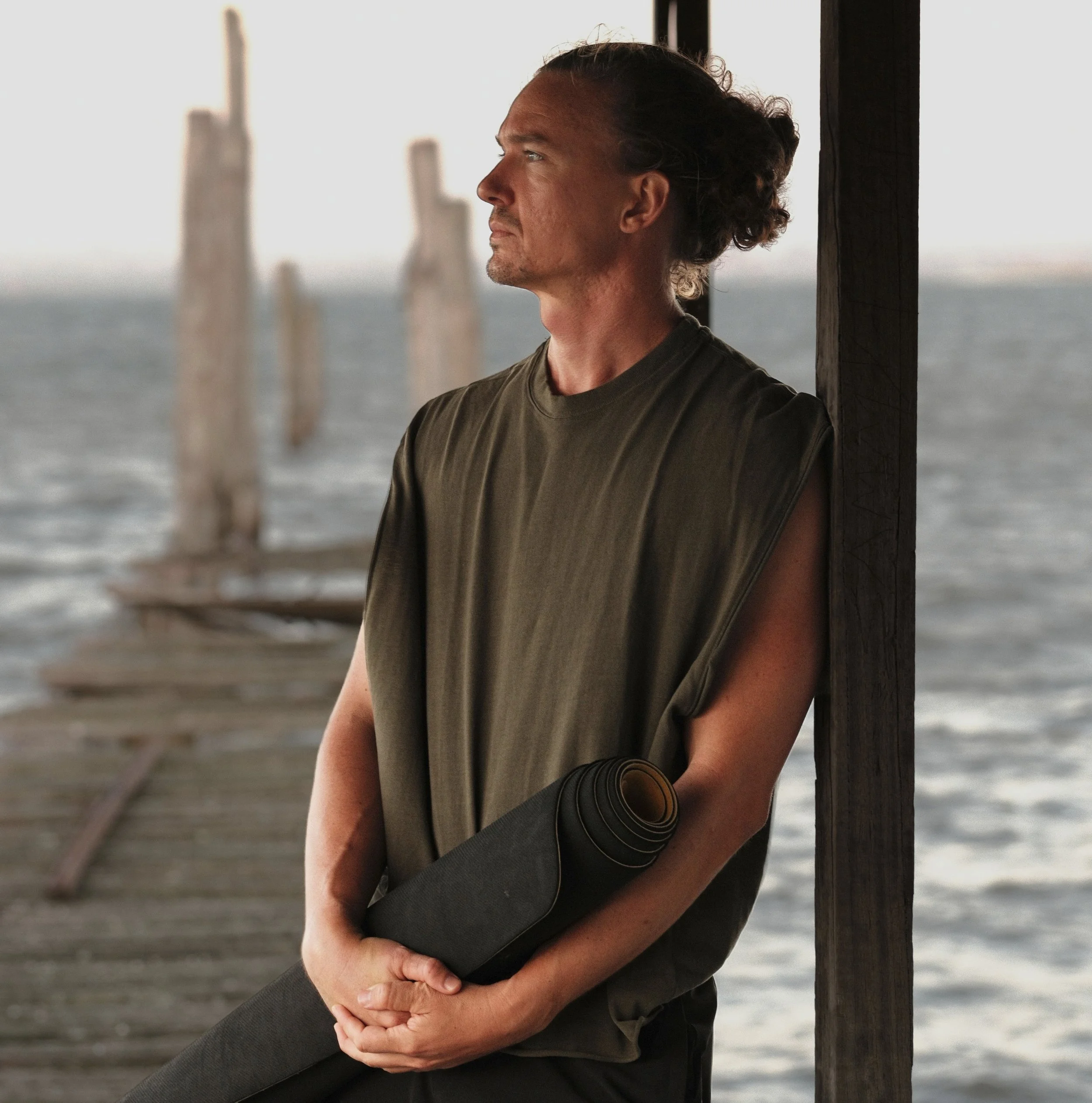 A male yoga teacher with long hair tied back, wearing a sleeveless olive green shirt, is standing by a wooden post on a dock by the water, holding a rolled-up yoga mat.