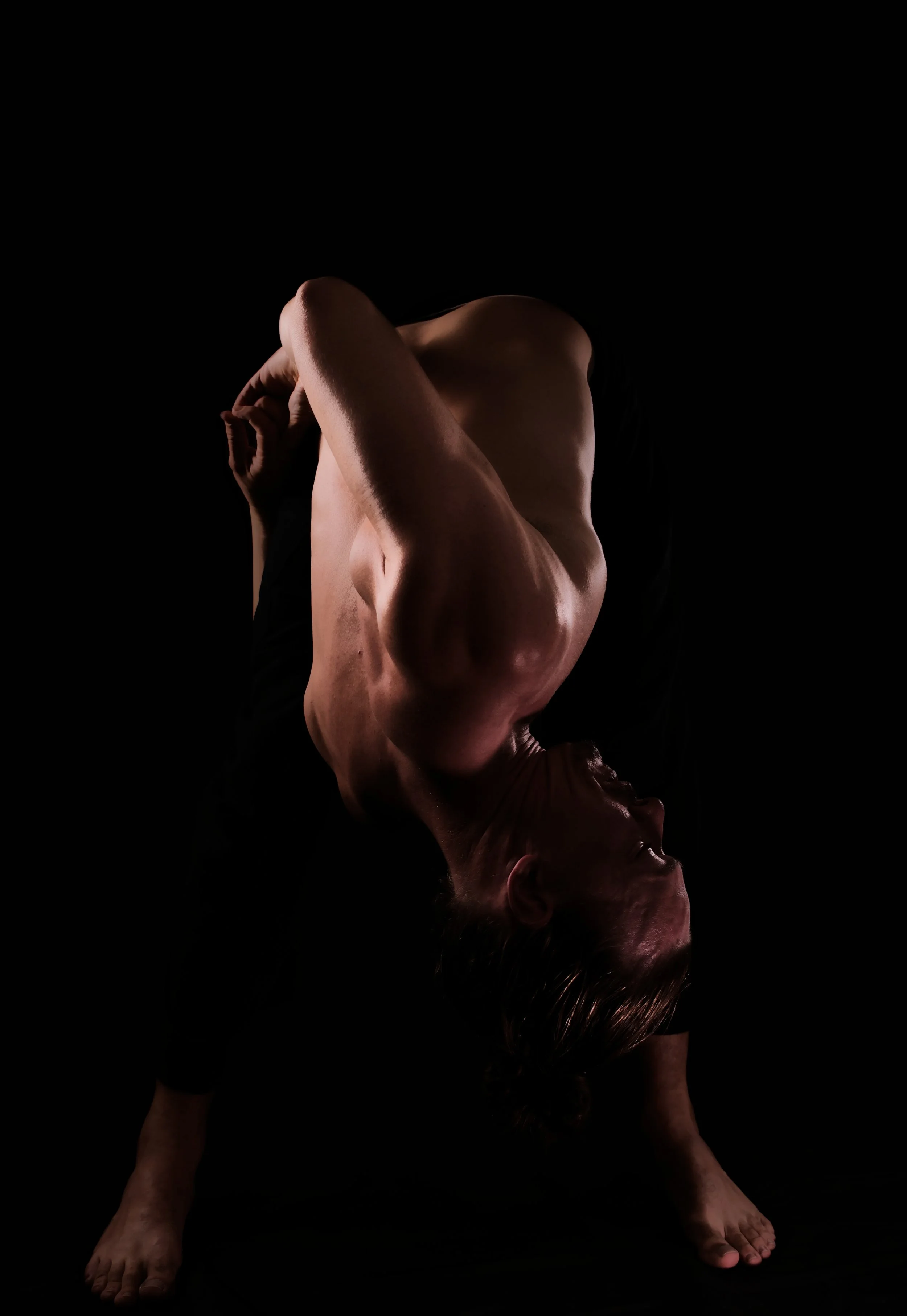 A person with tattoos performing a yoga pose against a black background, with one hand on the ground and the other arm bent across the chest.