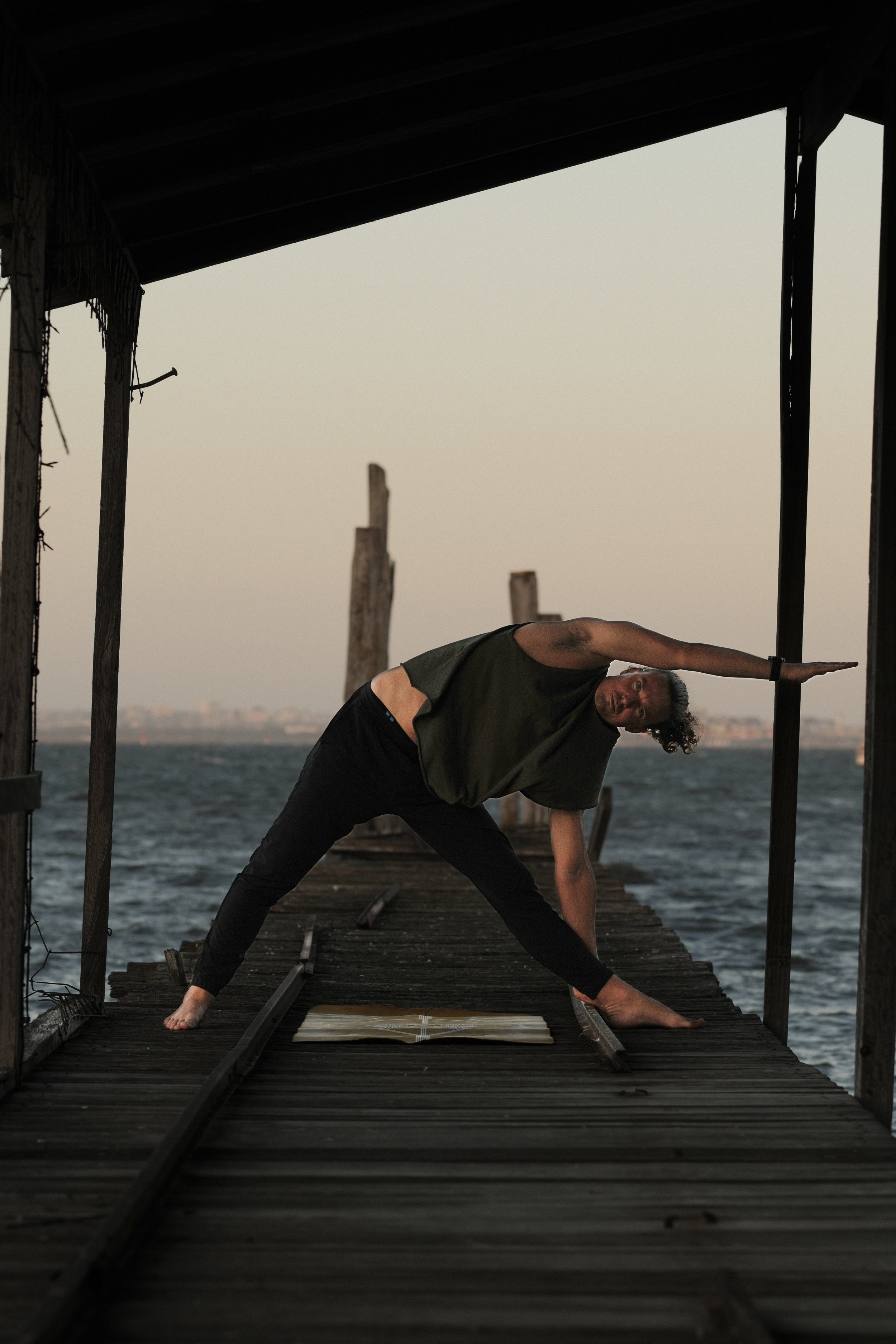 Person performing yoga stretch on a wooden pier by the water at sunset, with a book open in front of them.