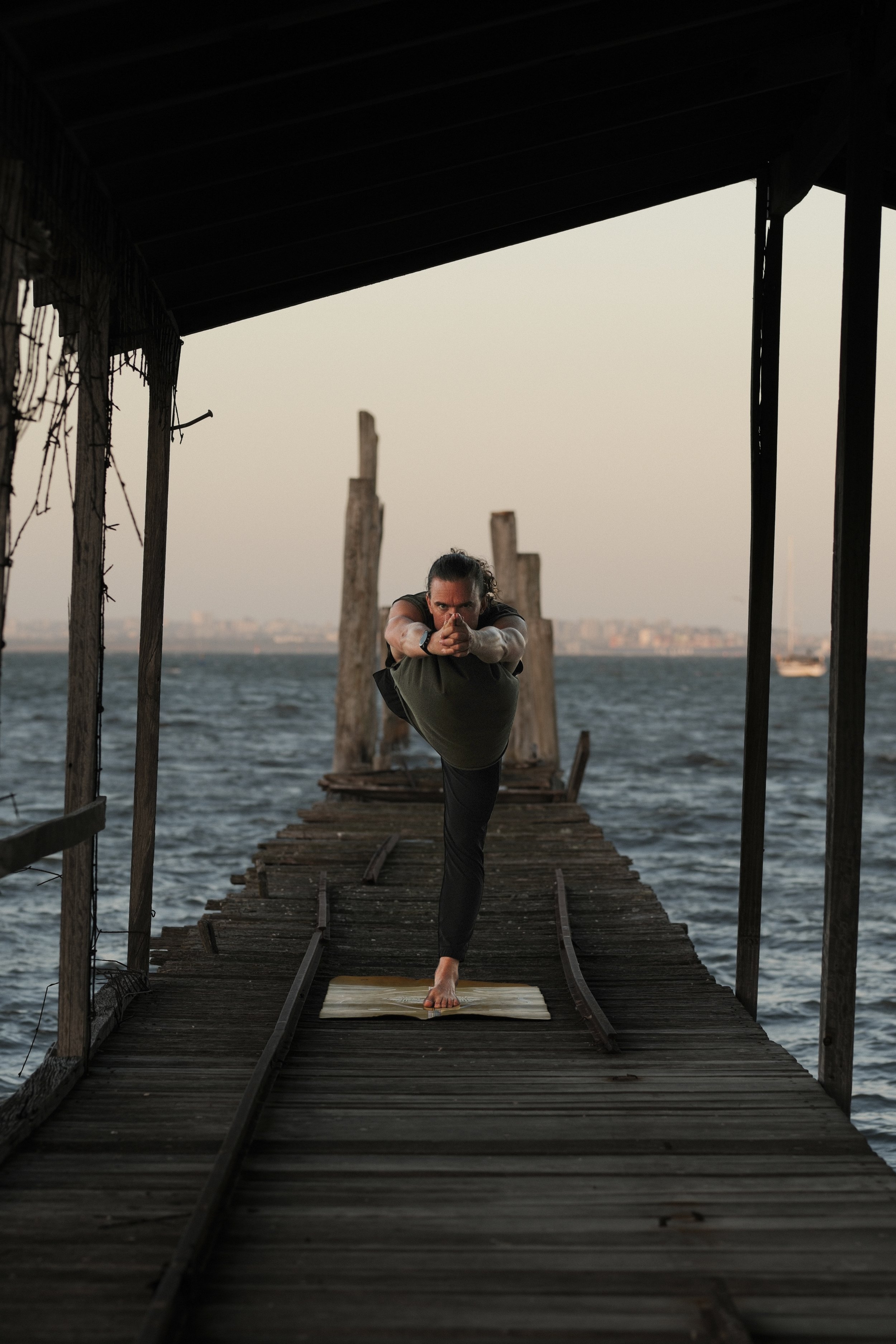 Person practicing yoga on a wooden dock by the water, performing a balancing pose with one leg lifted and arms extended forward.