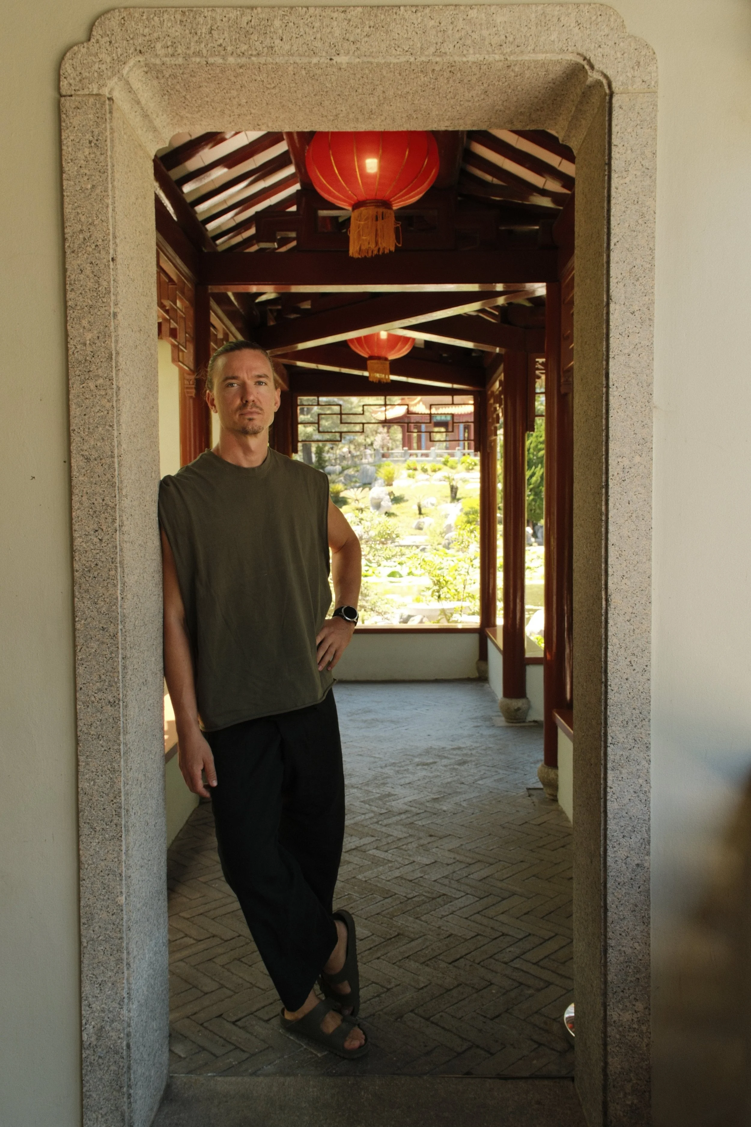 A man standing in a traditional Asian-style corridor with red lanterns and a lush garden outside, viewed through a stone doorway.