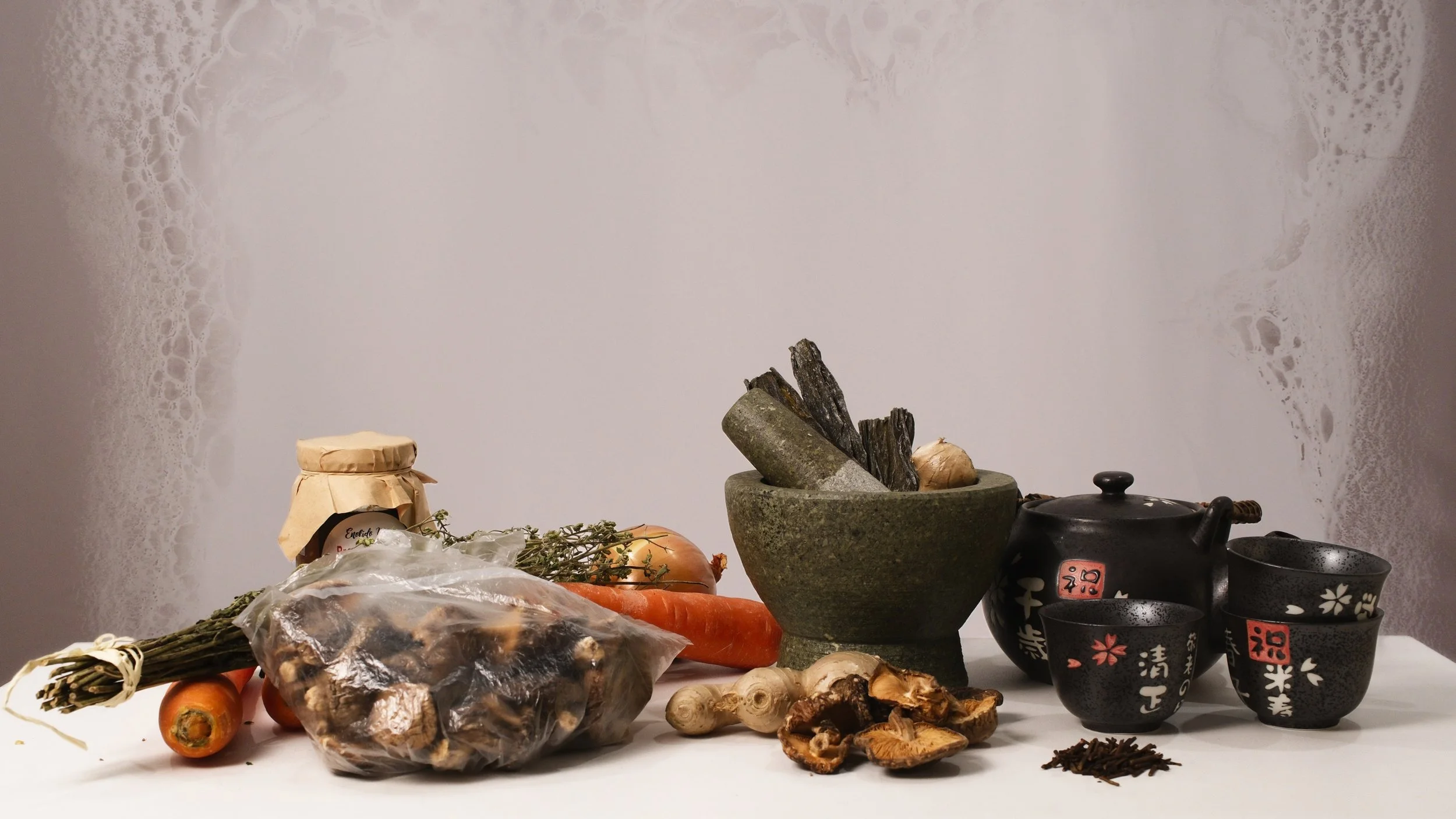A still life of fresh vegetables, dried herbs, a mortar and pestle with herbs, a traditional teapot and cups, and dried mushrooms on a white surface.