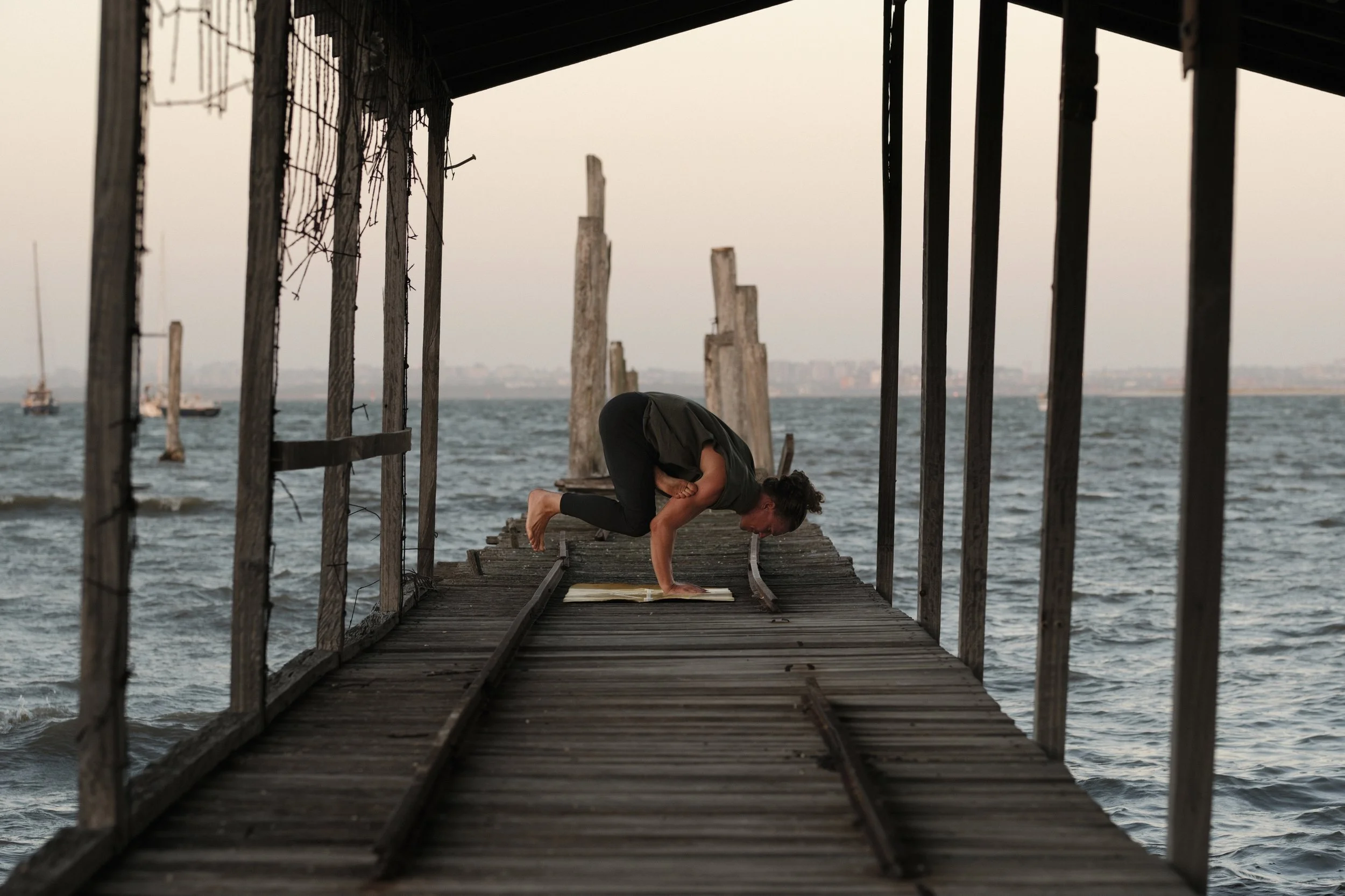 Person practicing yoga in a crab pose on a wooden pier over water, with boats and city skyline in the background.