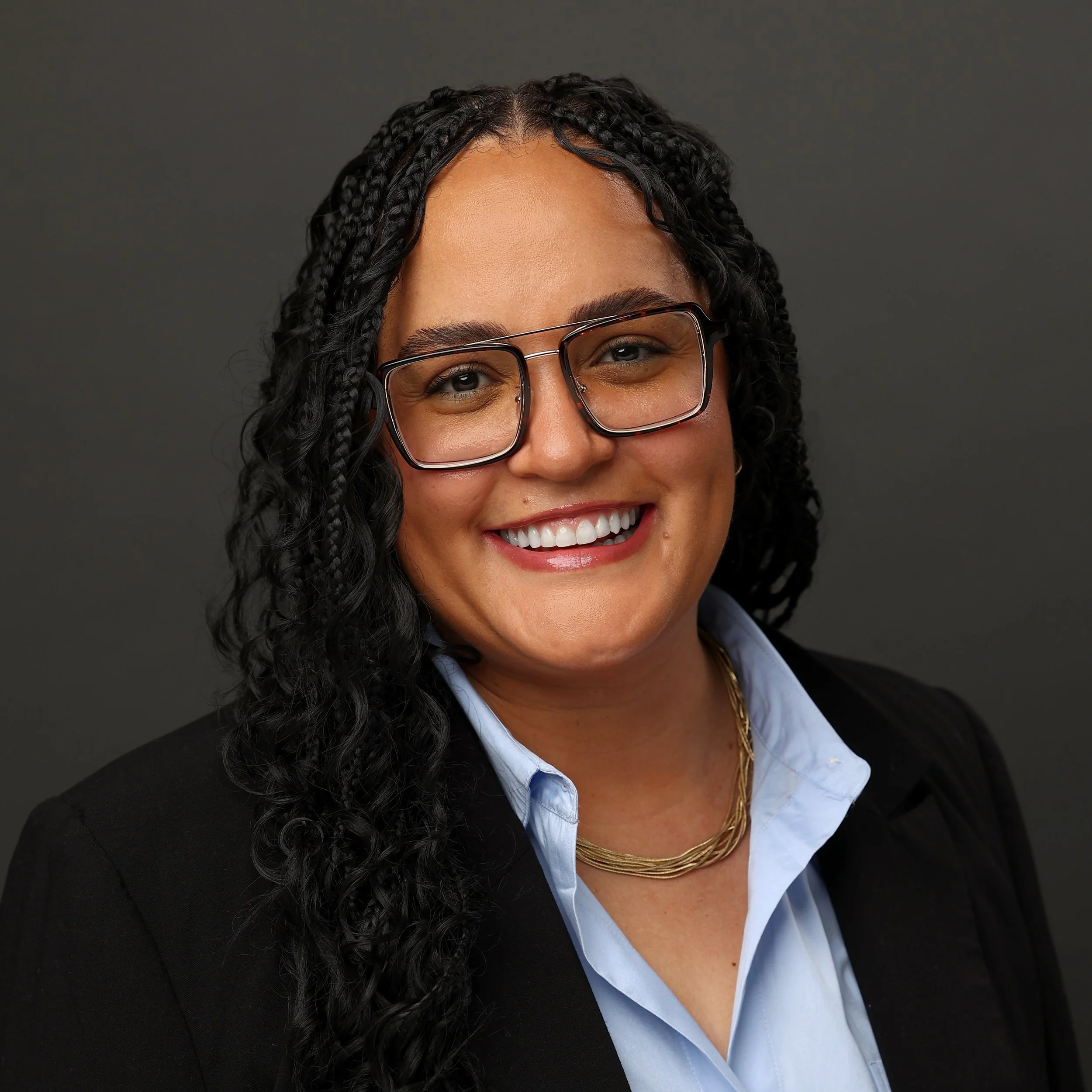 Close-up of a woman's face with glasses, smiling, with black braids, wearing a blue shirt and a gold necklace, against a dark gray background.