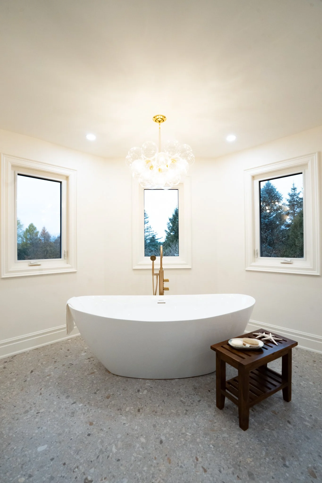 Bright bathroom with a standalone bathtub, three windows showing trees outside, a chandelier, and a small wooden table with bath products and starfish decoration.