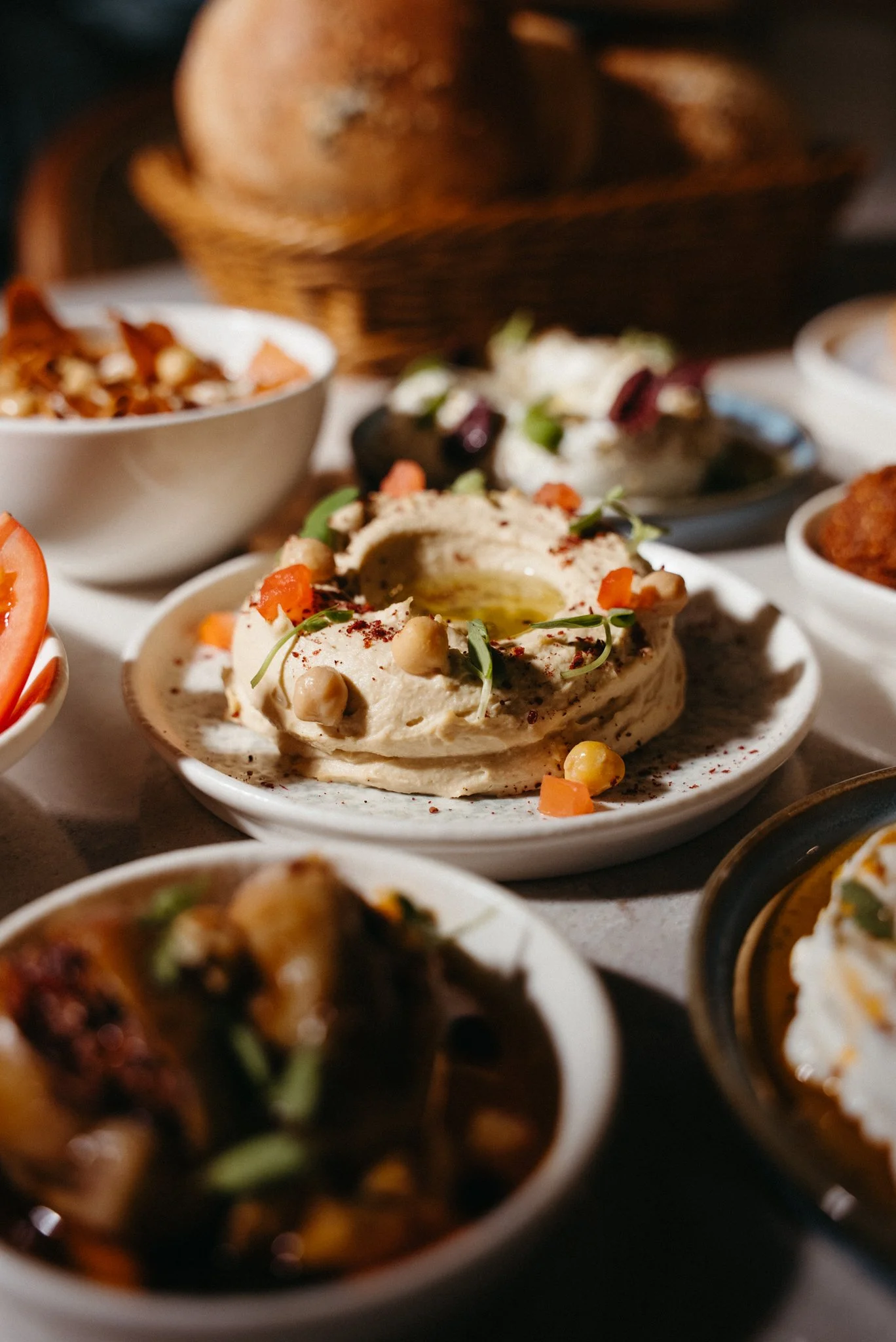 Various dishes of Middle Eastern food, including hummus garnished with chickpeas, vegetables, and spices on a white plate, surrounded by other bowls of appetizers and dips.