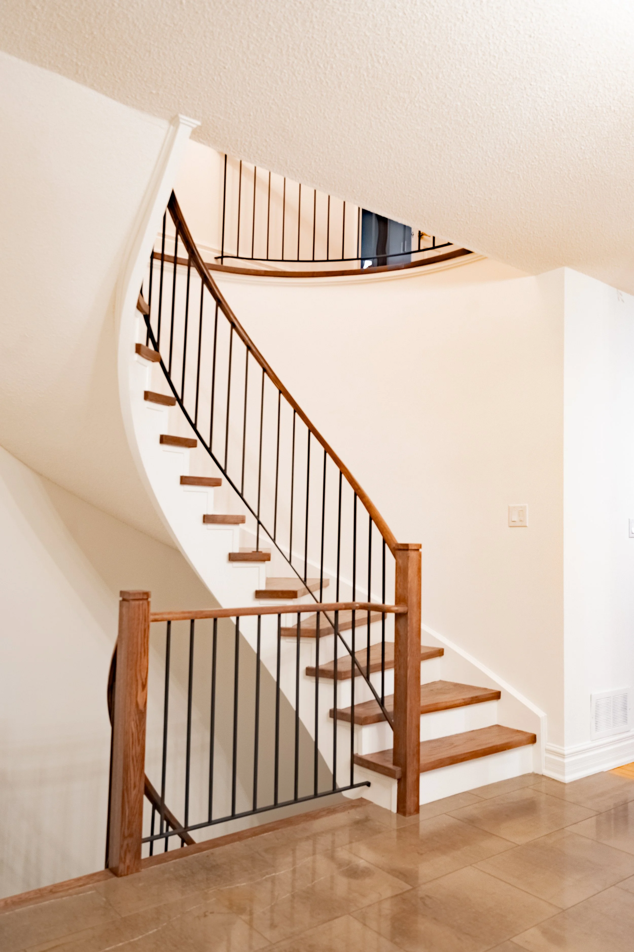 Interior view of a staircase with wooden steps, black metal railing, and a curved design, leading to an upper floor in a home with white walls and brown hardwood flooring.