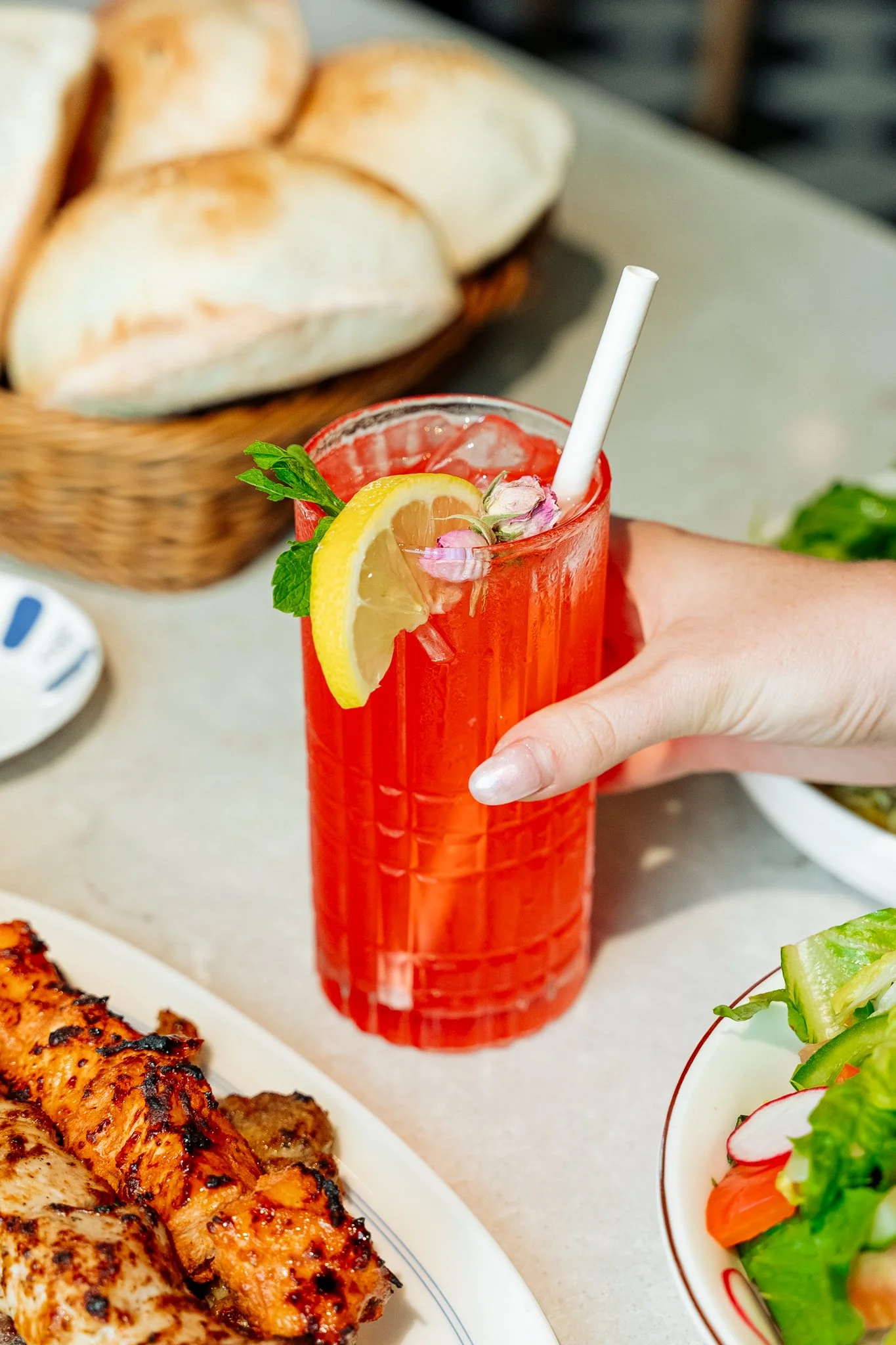 A hand holding a tall glass of pink drink garnished with lemon, mint, and flower petals, with bread in a basket and grilled chicken on a plate in the background.