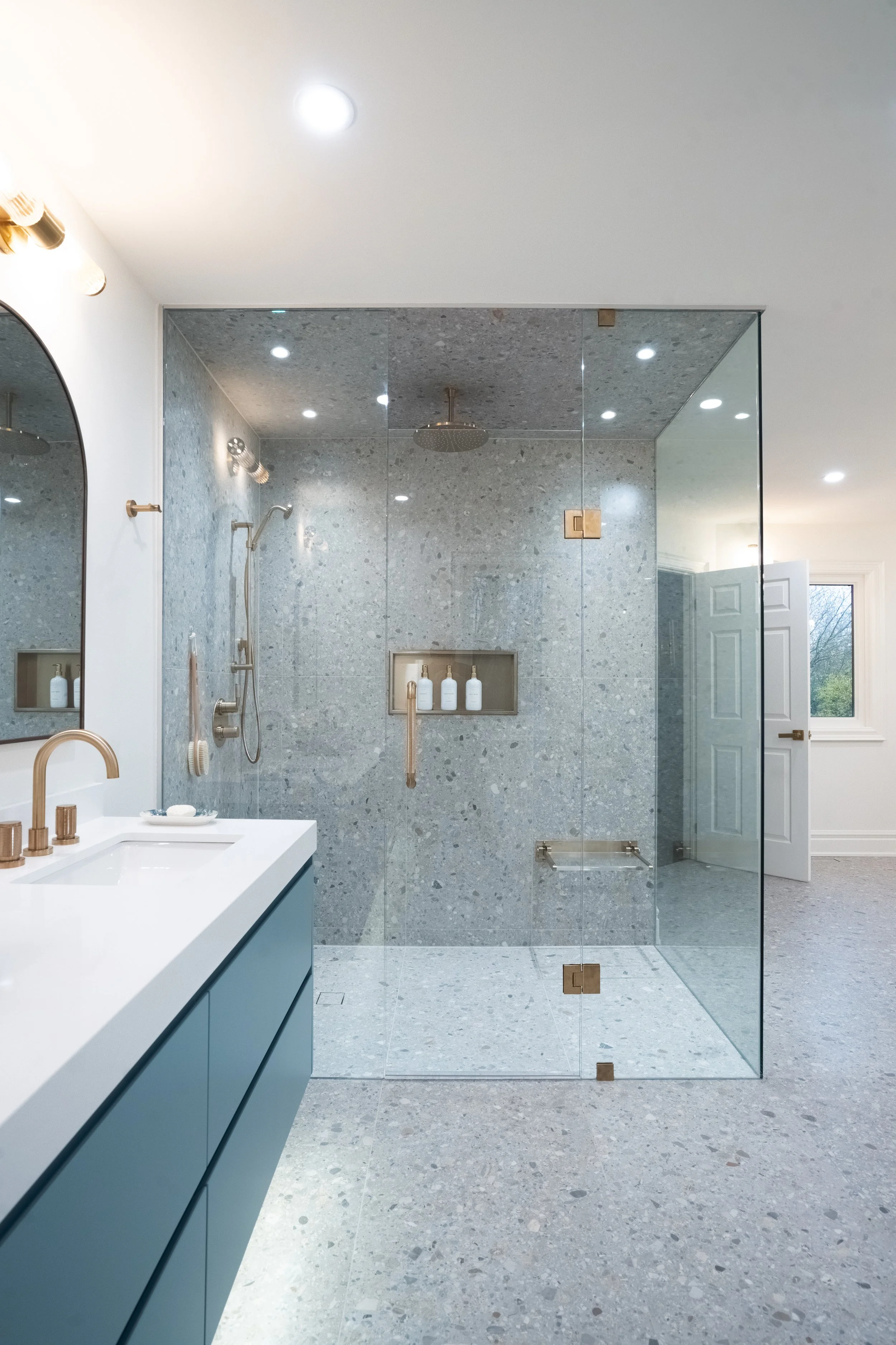 Modern bathroom with a glass-enclosed shower area with gray textured tiles, a built-in shelf with bottles, and a white vanity with a blue cabinet and gold fixtures.