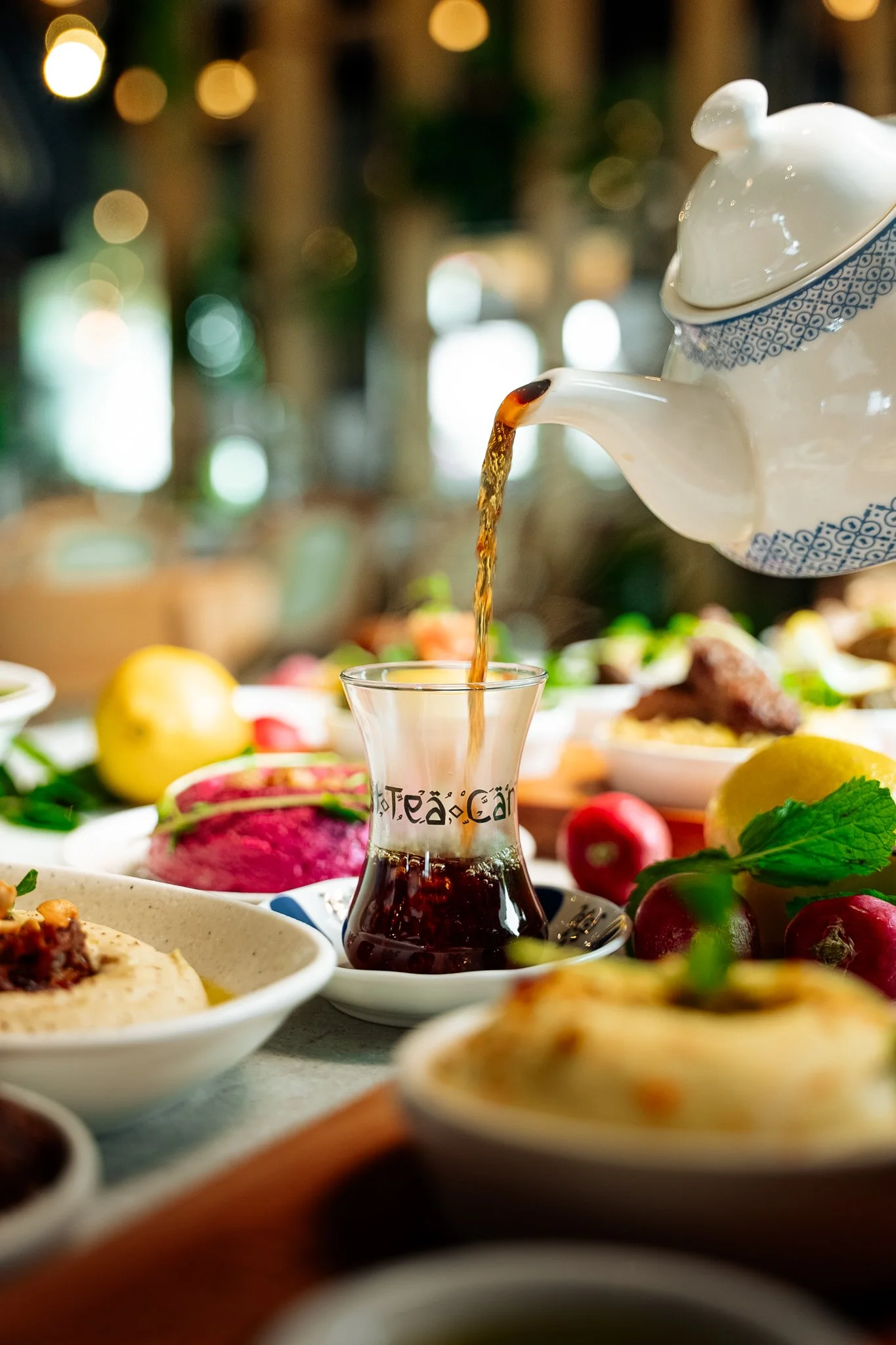 Tea being poured from a teapot into a small glass on a table with various dishes and fruits, including radishes, lemons, and other food items, in a cozy indoor setting.
