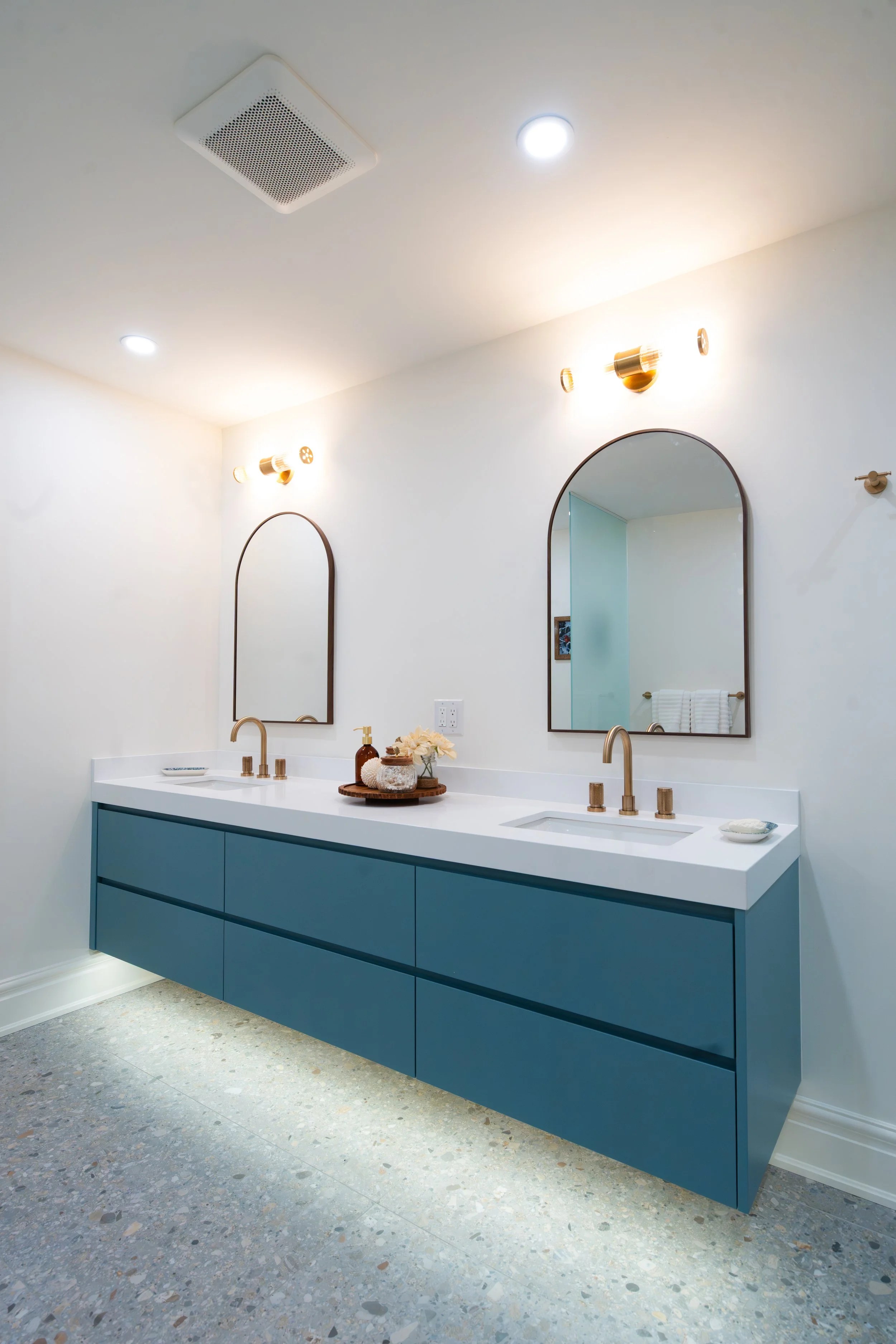 Modern bathroom vanity with two sinks, gold fixtures, two arched mirrors, and decorative items including soap dispenser and flowers.