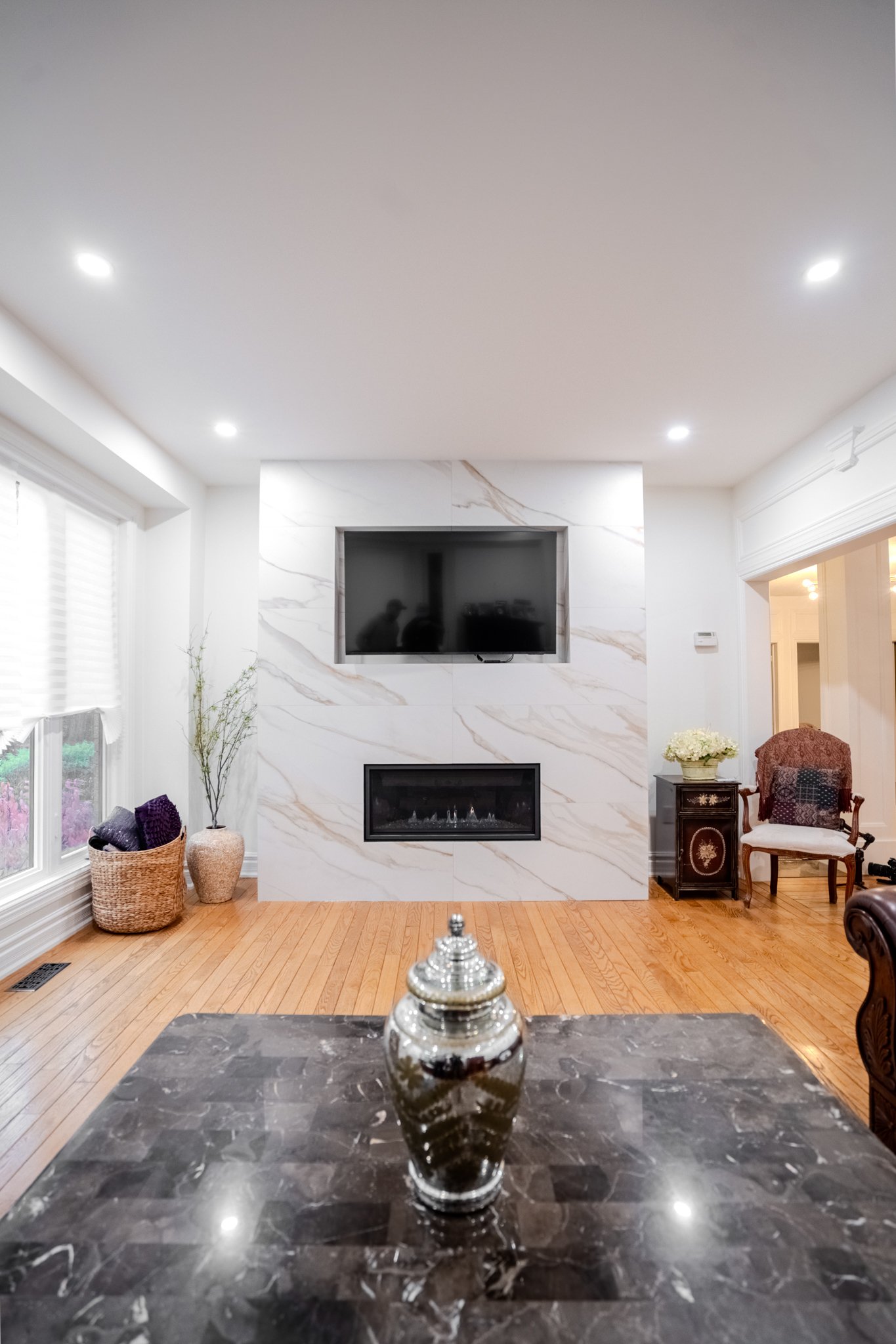 Living room with a wall-mounted TV above a fireplace, wooden floor, a vase with tall branches, basket with purple cushions, upholstered armchair with floral cushion, and a small side table with flowers.