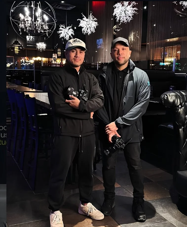 Two men in dark jackets and caps standing inside a decorated restaurant or club, each holding a camera, with hanging snowflake decorations and chandeliers overhead.