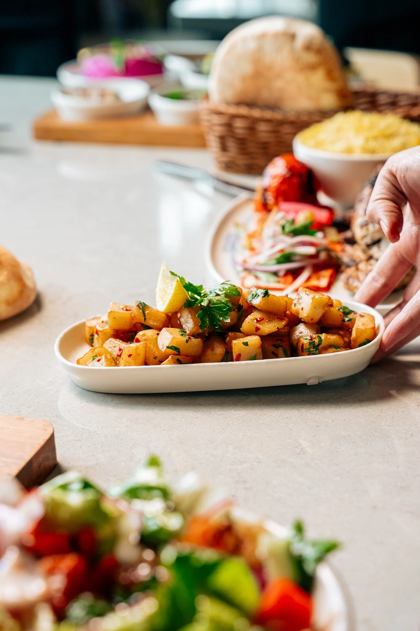 A white oval dish with chopped seasoned potatoes garnished with cilantro and a lemon wedge, on a light-colored table with various baked goods and dishes in the background.