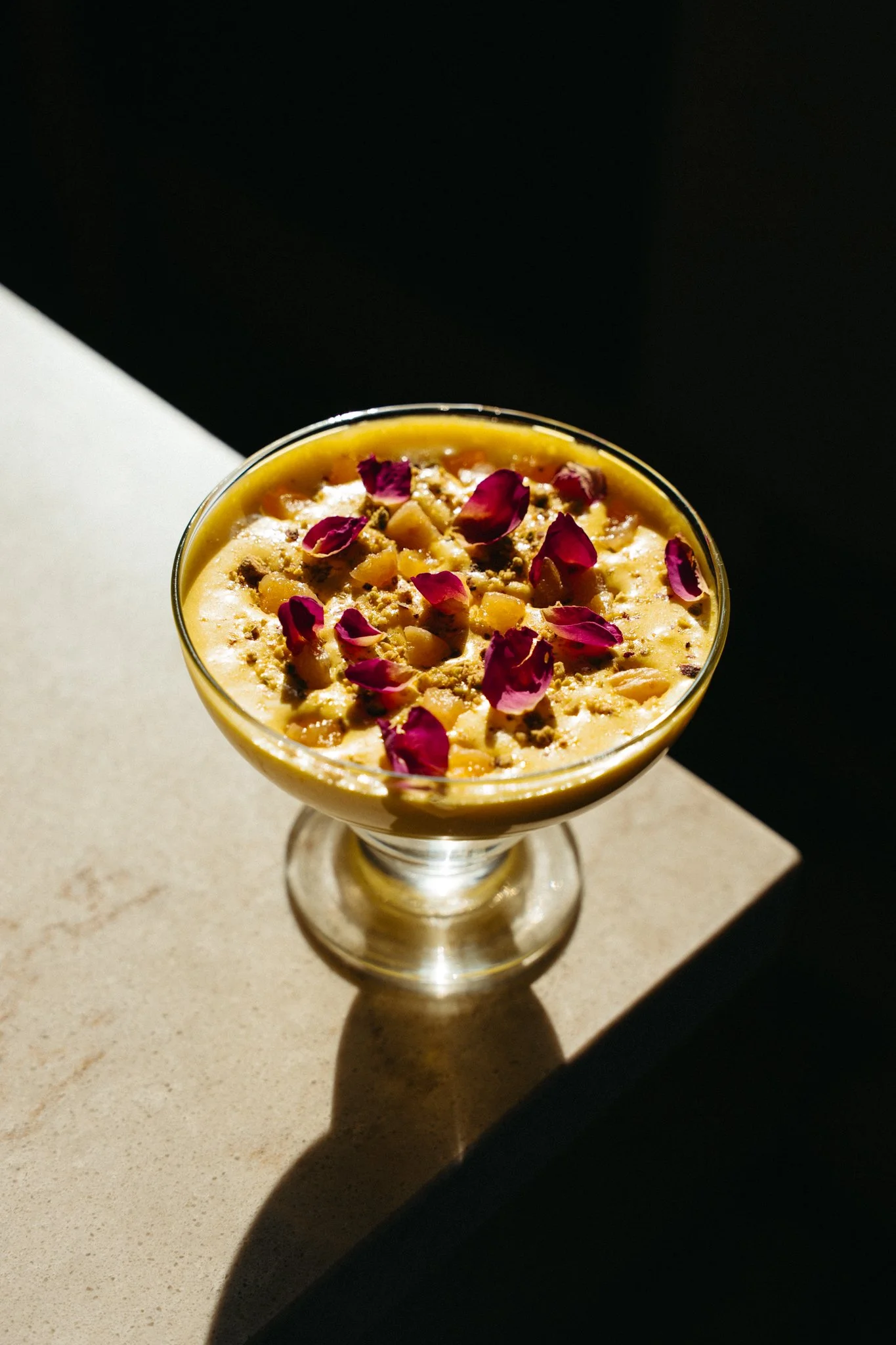 A glass dessert cup filled with a yellow pudding topped with pink rose petals and crushed nuts, placed on a beige surface with shadows and a dark background.