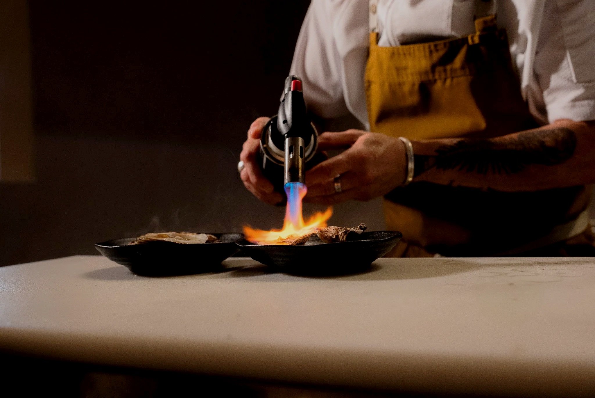 Assorted small appetizers on a wooden platter, including mini sandwiches, crackers with toppings, and skewered bites, with glasses in the background.
