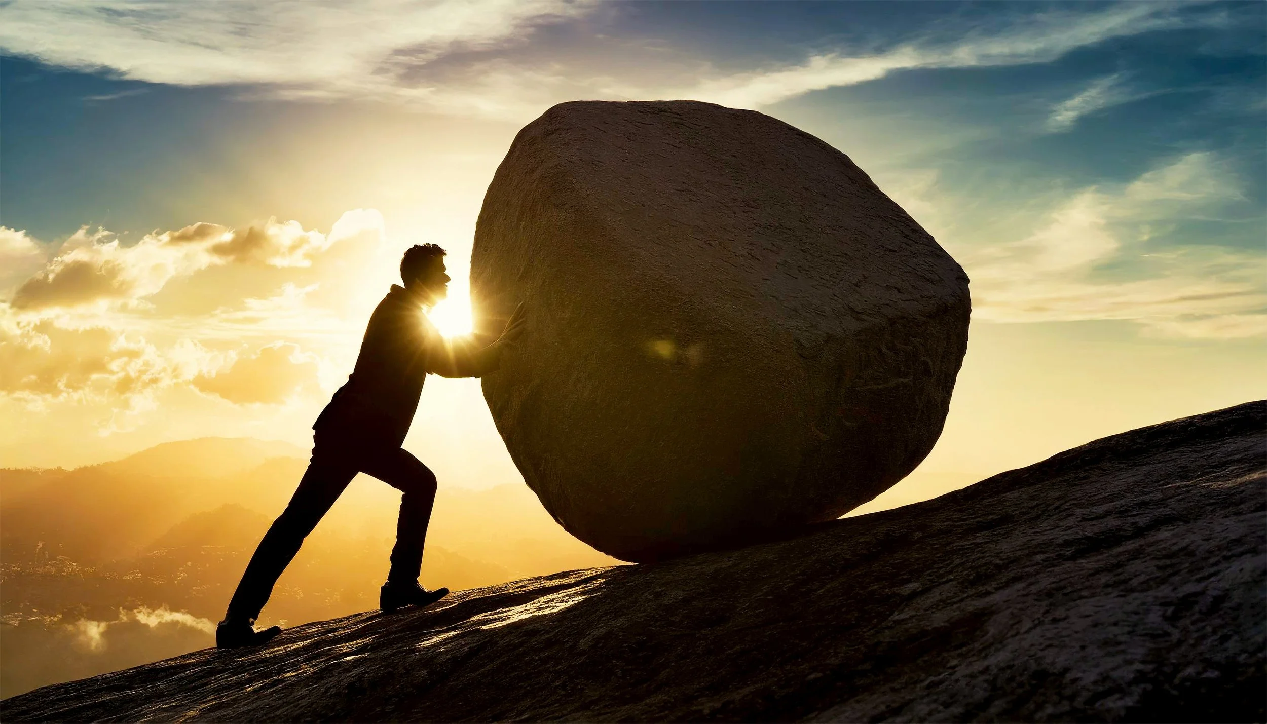 Silhouette of a person pushing a large boulder uphill against a sunset sky with clouds.