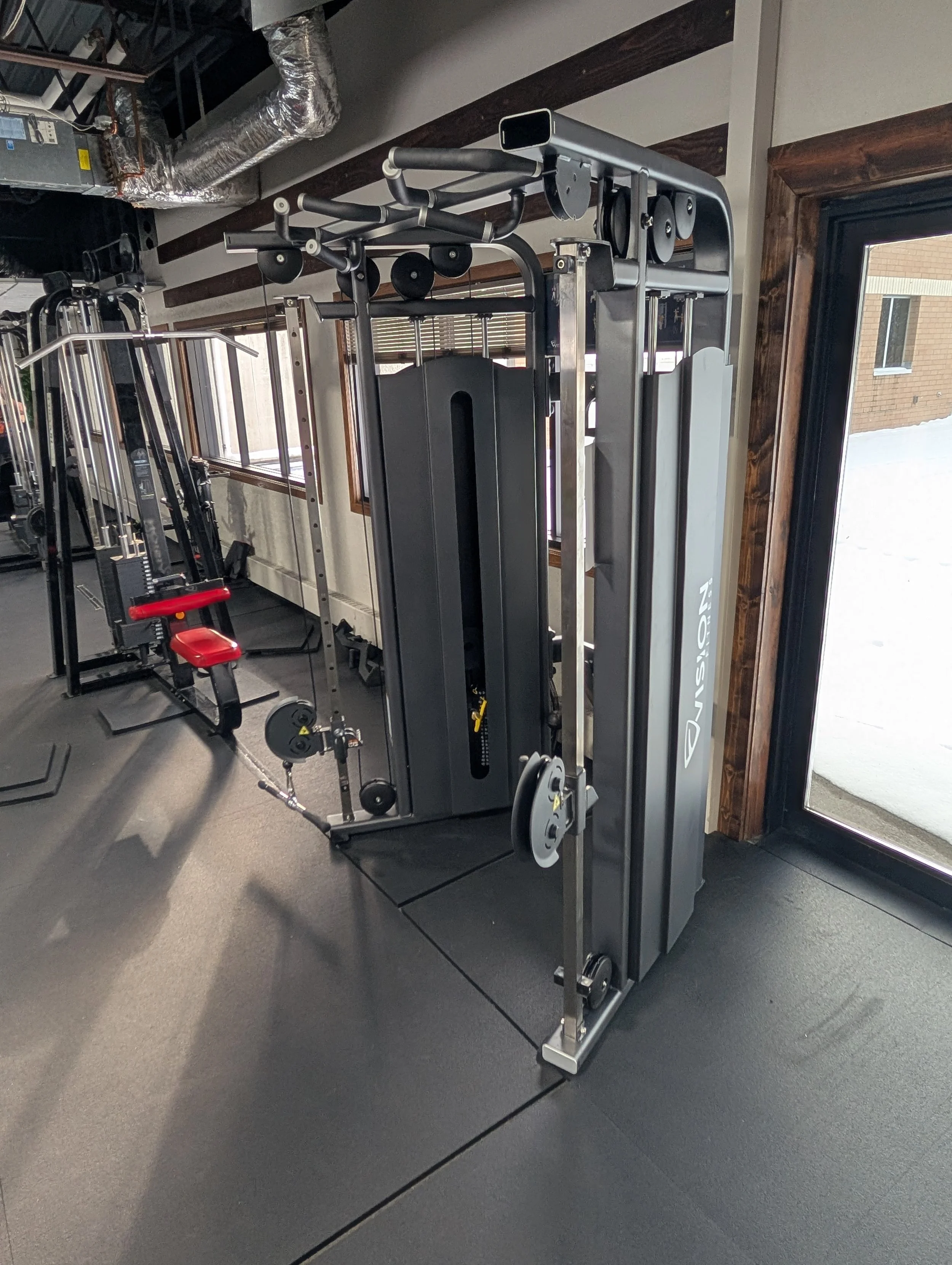 Fitness gym equipment including a cable machine with weight stacks and pulleys, a seated chest press with red padding, and other workout machines in a well-lit room with windows and wooden accents.