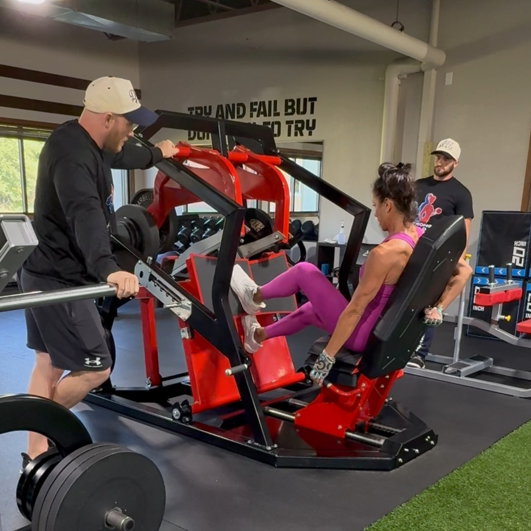 A woman in pink workout clothes is performing leg presses on a specialized gym machine with two men assisting her, inside a gym.