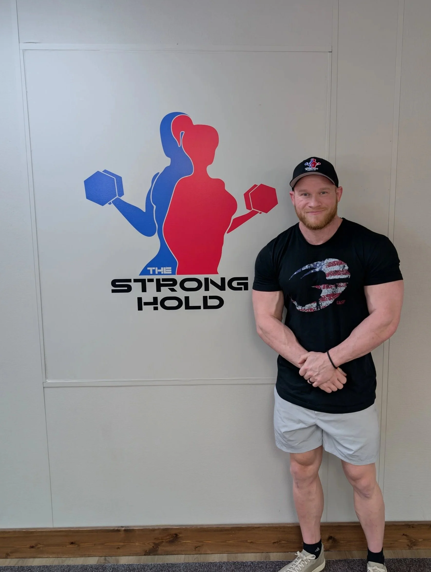 A man standing in front of a gym wall with a logo that features a silhouette of a woman and a man lifting weights, with the words 'The Strong Hold' underneath.