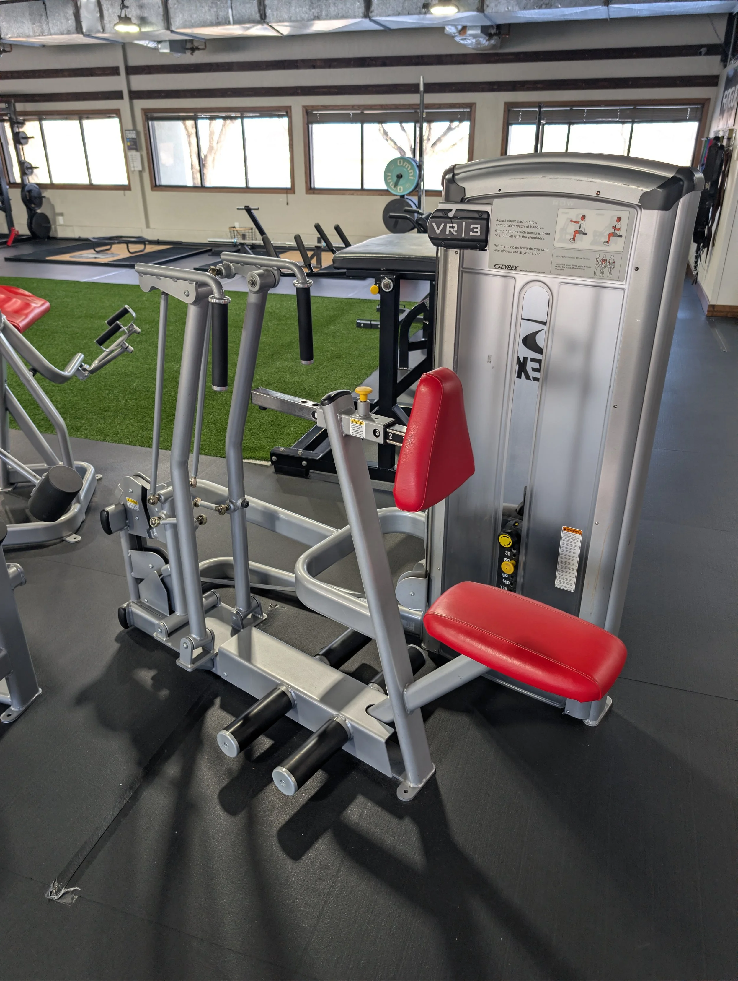 Red and silver seated exercise machine in a gym with rubber flooring, windows in the background, and other fitness equipment.