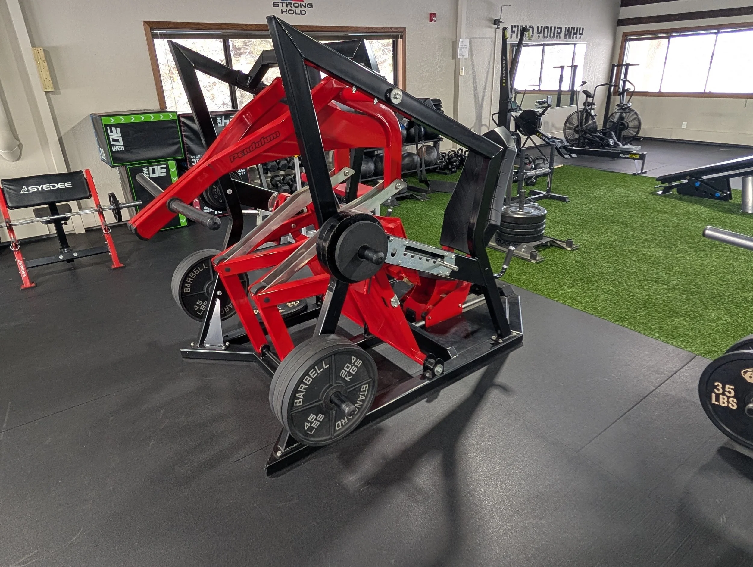 A gym with various equipment, including a red and black strength machine in the foreground, weight plates, a rowing machine, and a green turf area with a bench, and large windows in the background.