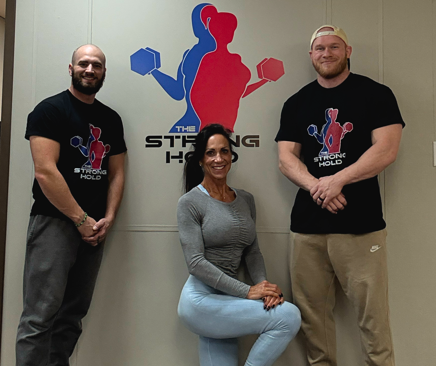 Three people posing indoors in front of a wall with a fitness logo, two men standing on either side and a woman sitting in the middle. The wall features a logo of two stylized human figures lifting dumbbells, one blue and one red, with the text 'The Strong Hold'.