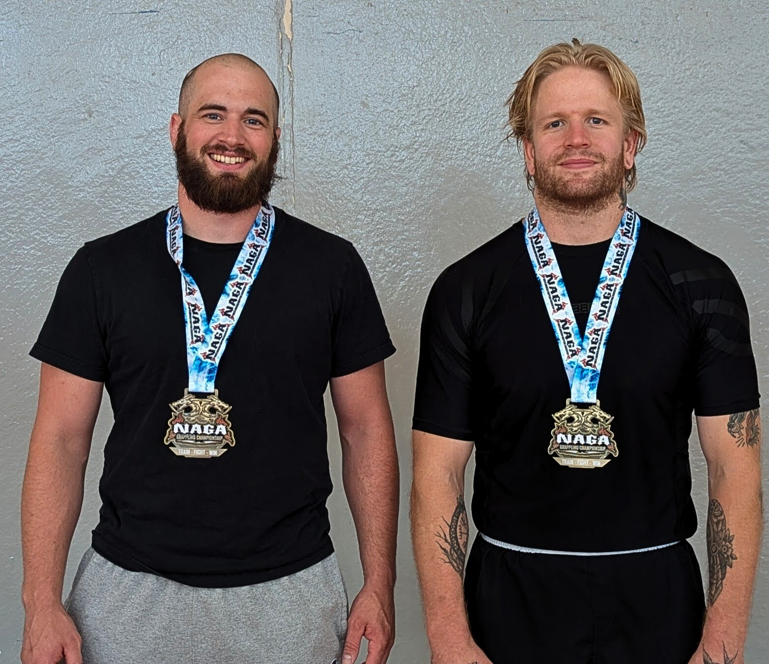 Two men standing side by side wearing medals around their necks with a "NAGA" logo, dressed in black athletic shirts, against a light gray textured wall.
