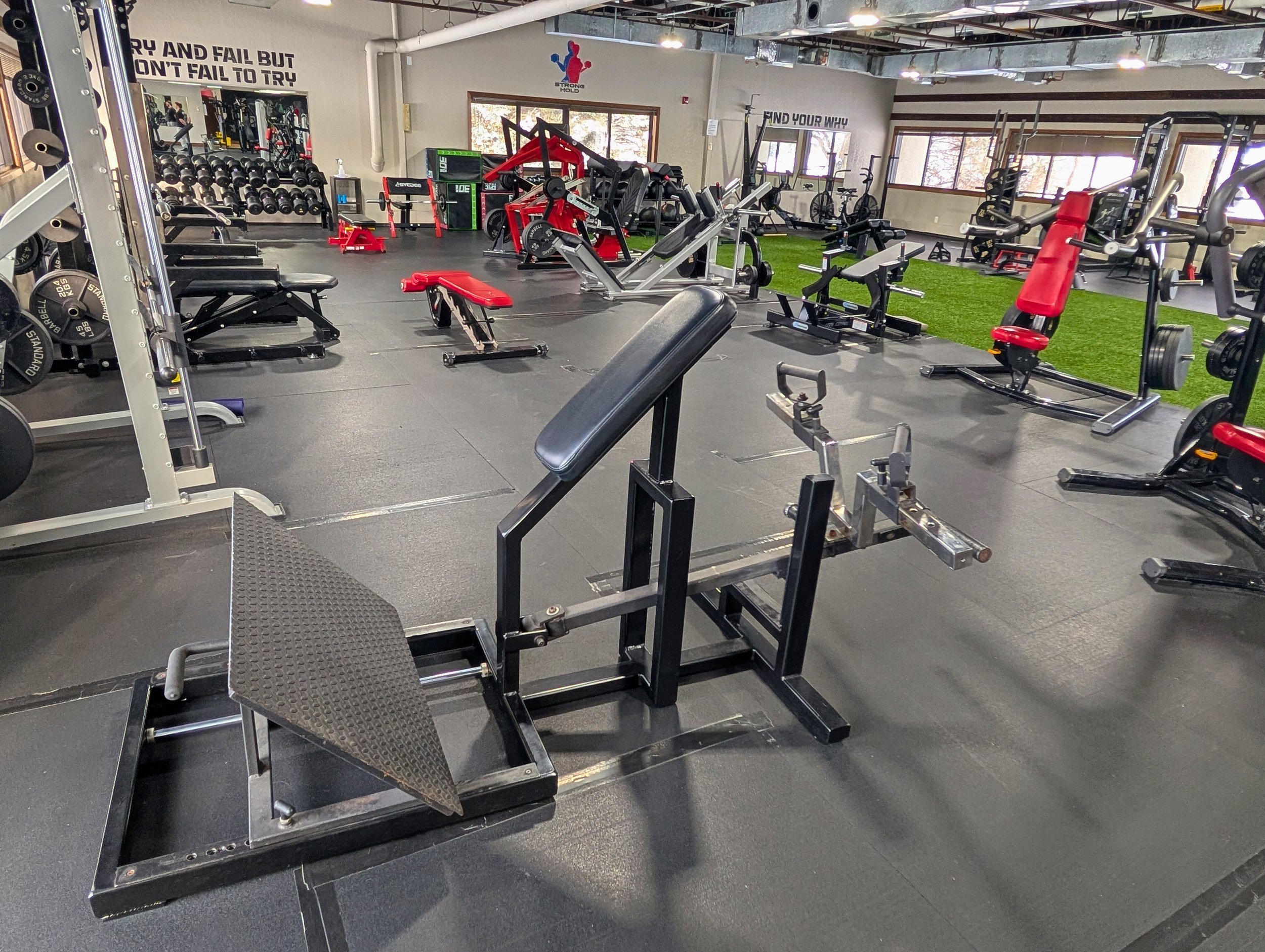 Empty gym with various workout equipment including weight benches, dumbbells, cardio machines, and a turf area, with motivational quotes on the walls.
