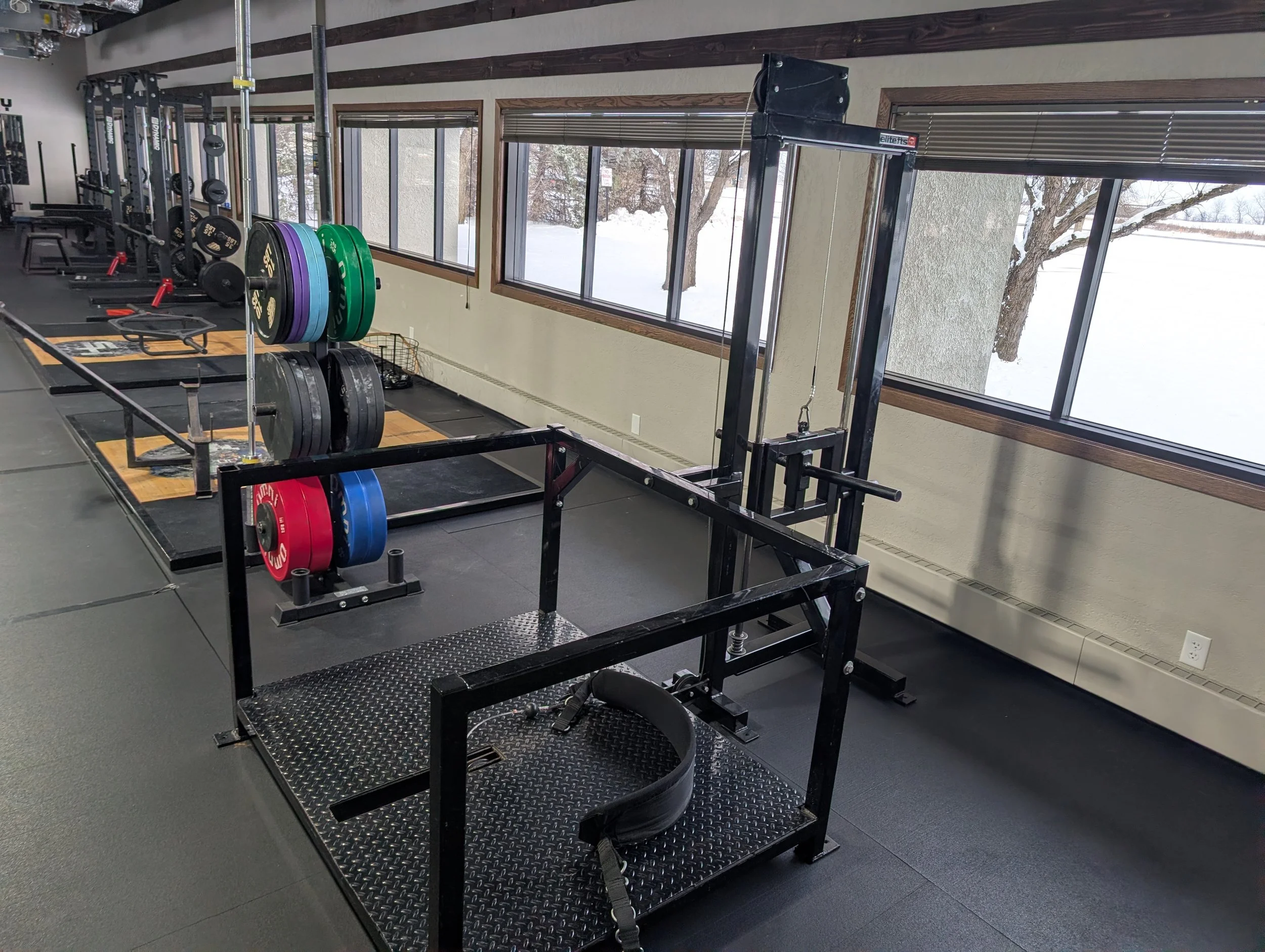 Indoor gym with weightlifting equipment, including weight plates, barbells, and a squat rack, next to large windows showing snow outside.
