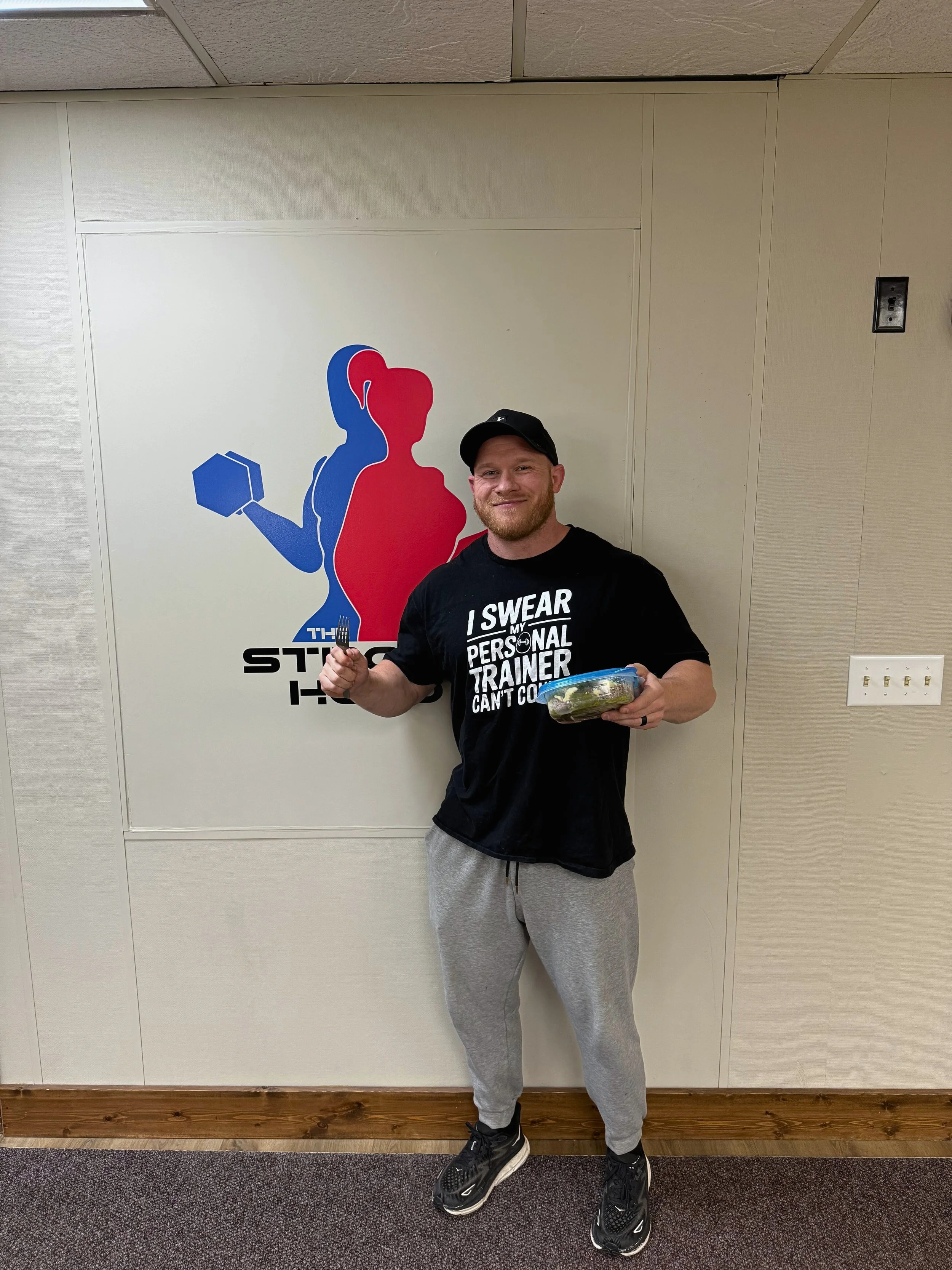 A man standing in front of a sports-themed wall mural holding a container of salad and a fork, wearing a black cap, black T-shirt, and gray sweatpants, smiling at the camera.