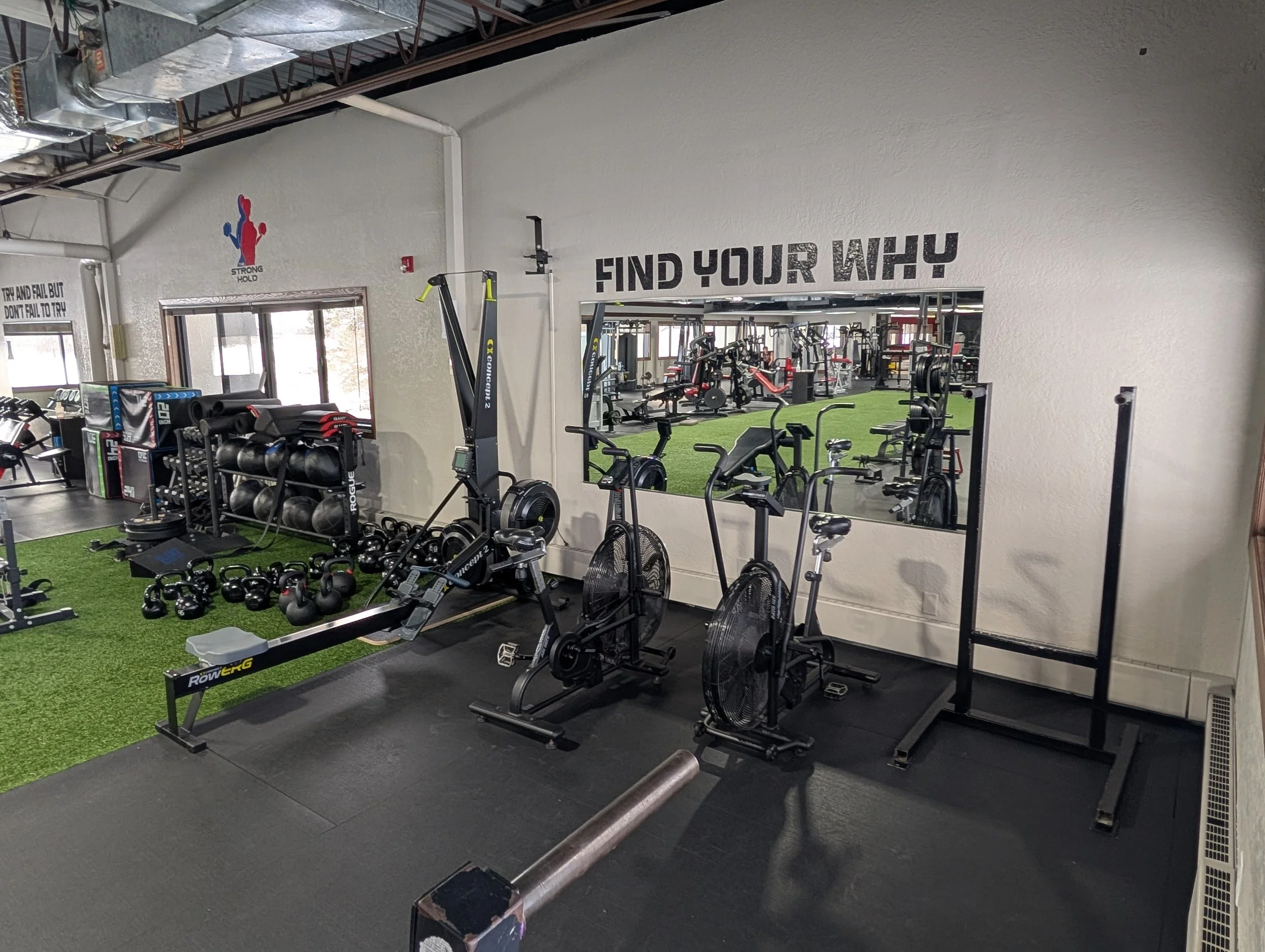 A gym with various workout equipment including kettlebells, rowing machines, and stationary bikes. A large mirror on the wall reflects the gym interior, with motivational text 'FIND YOUR WHY' above it.