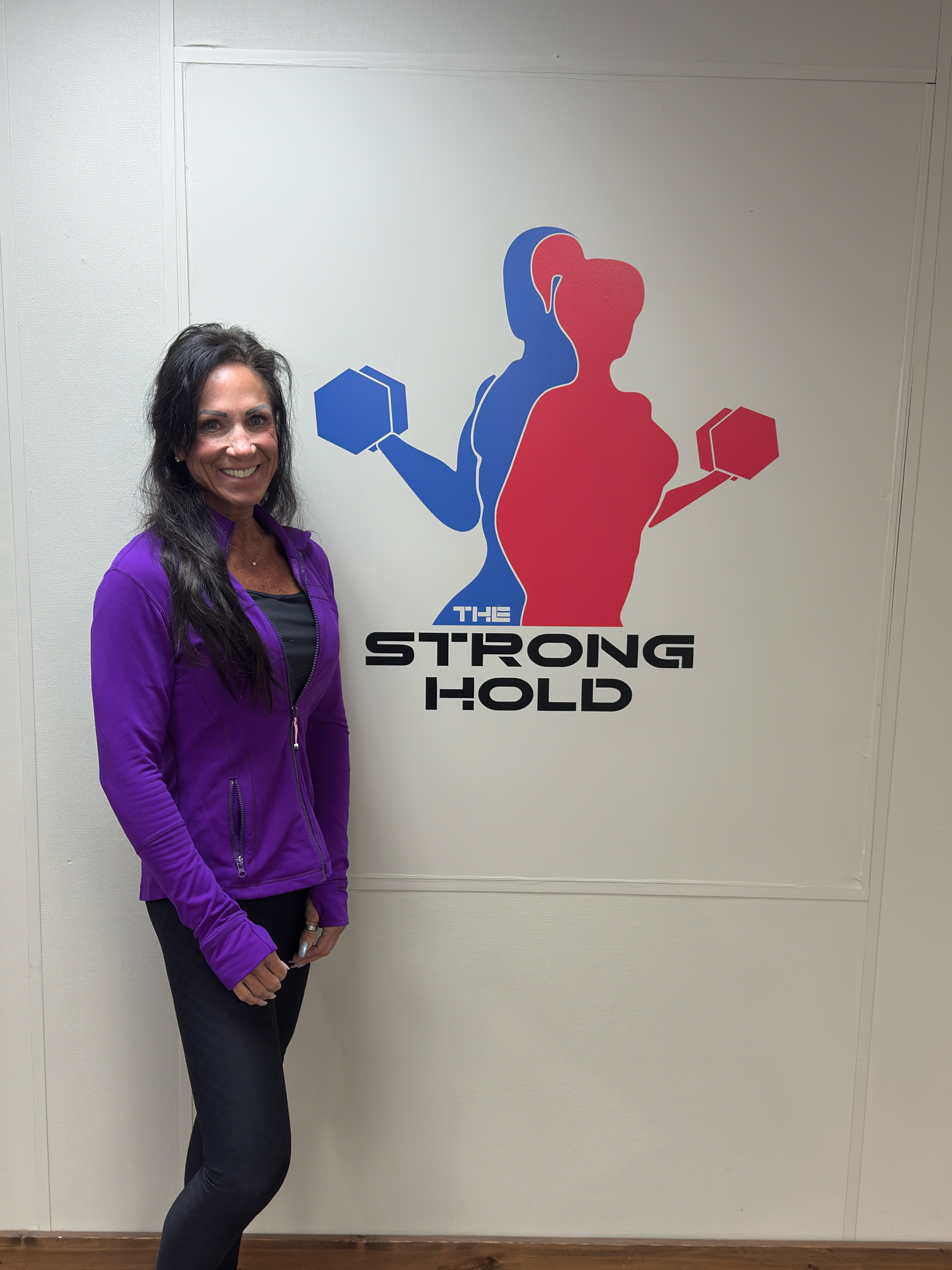Woman with dark hair wearing a purple jacket and black pants standing next to a sign of two female silhouettes lifting dumbbells with the words "The Strong Hold" underneath.