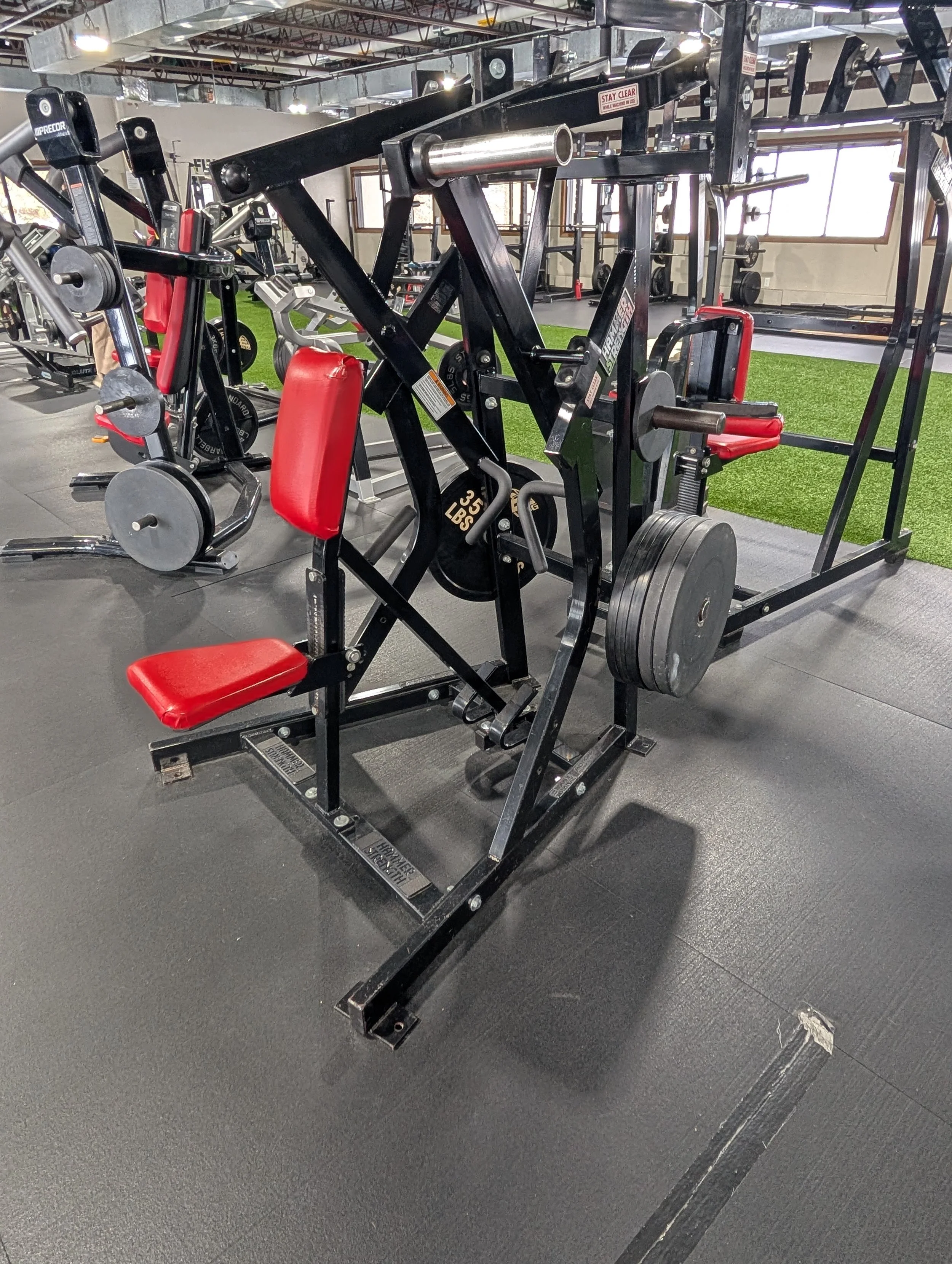 A gym with various workout equipment including a black and red seated leg press machine with weight plates, a cardio machine, and a squat rack, all on a rubberized floor with a green turf area and large windows in the background.