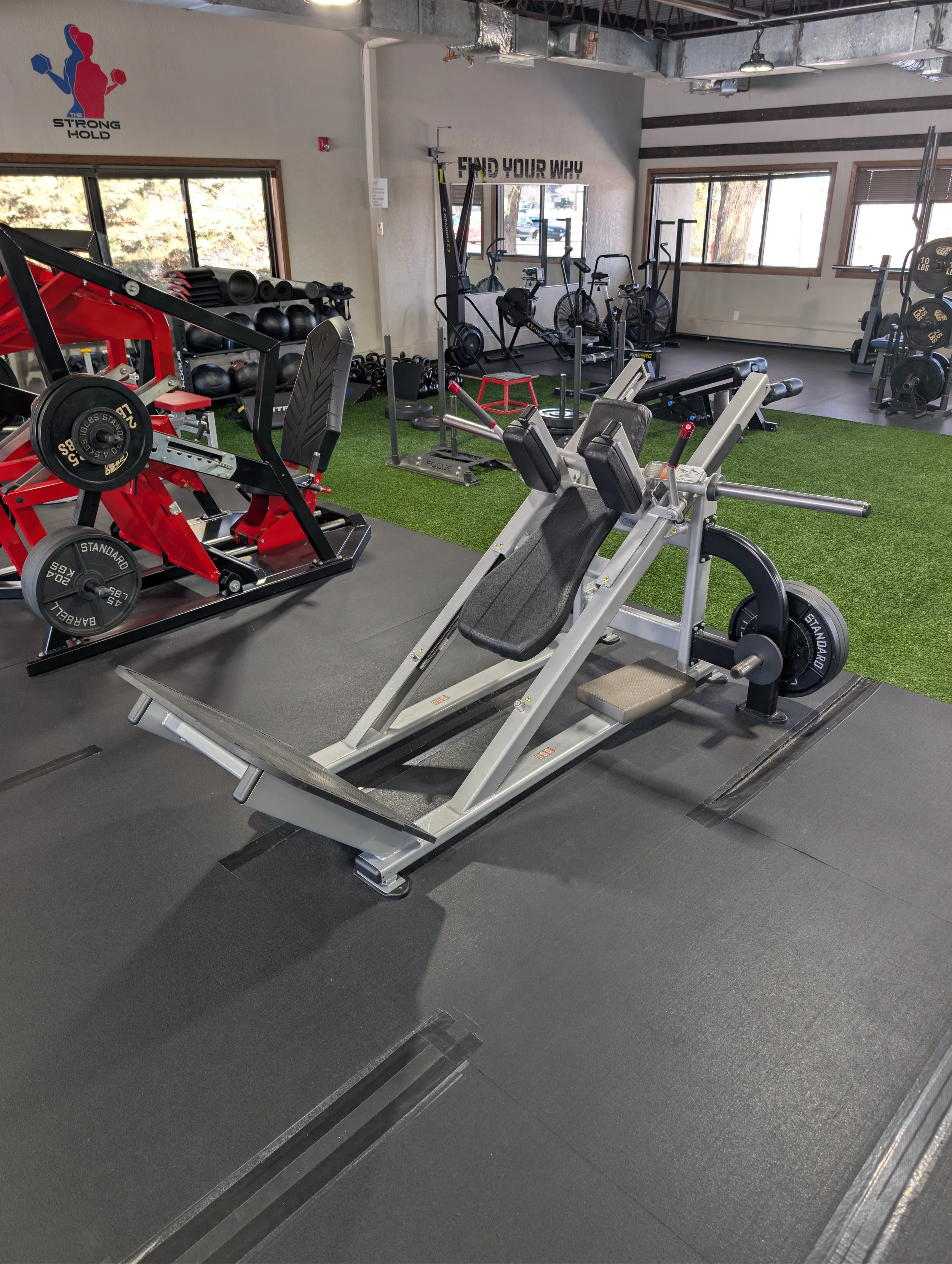 Gym equipment including a leg press machine in the foreground, with various weight machines, cardio bikes, and free weights in the background, along with motivational signs on the wall.