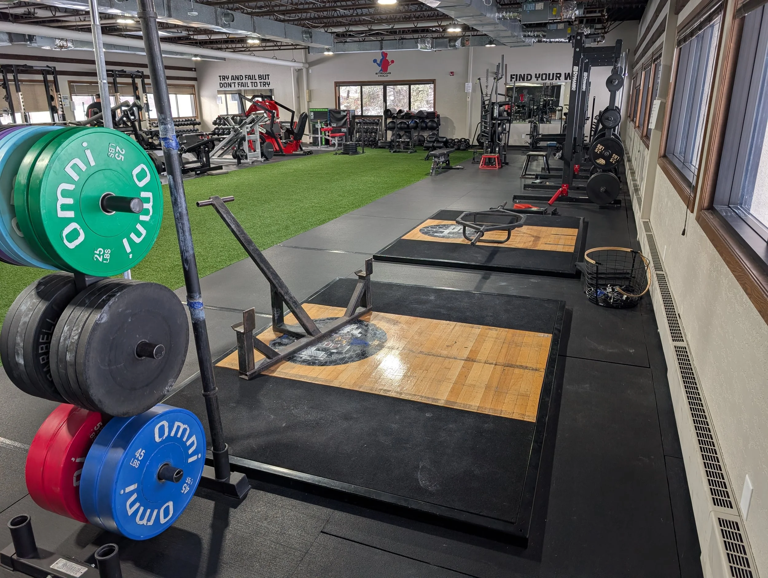 Indoor gym with weightlifting equipment, platforms, and exercise machines, with a green turf section in the background and motivational signs on the walls.