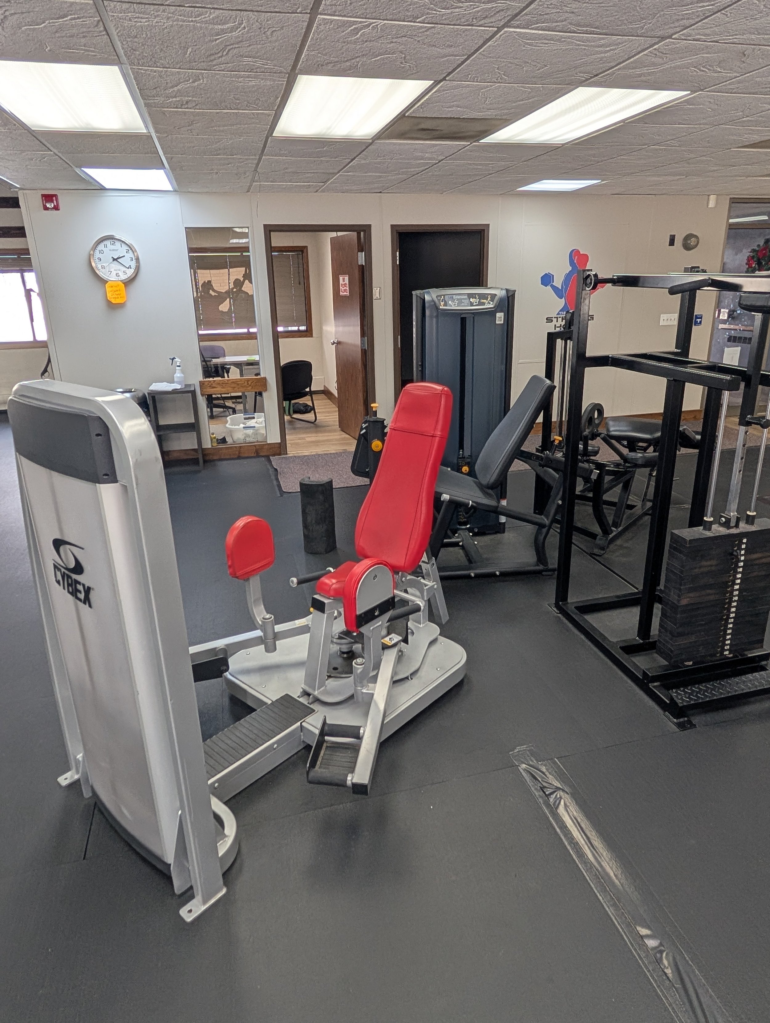 A gym room with various exercise equipment, including a Cybex leg press machine, a red and black exercise chair, and a sled with weights. The room has a gray rubber floor, a drop ceiling with fluorescent lights, and a clock on the wall showing 10:35. In the background, there's a small office with a chair and a table, visible through an open doorway.