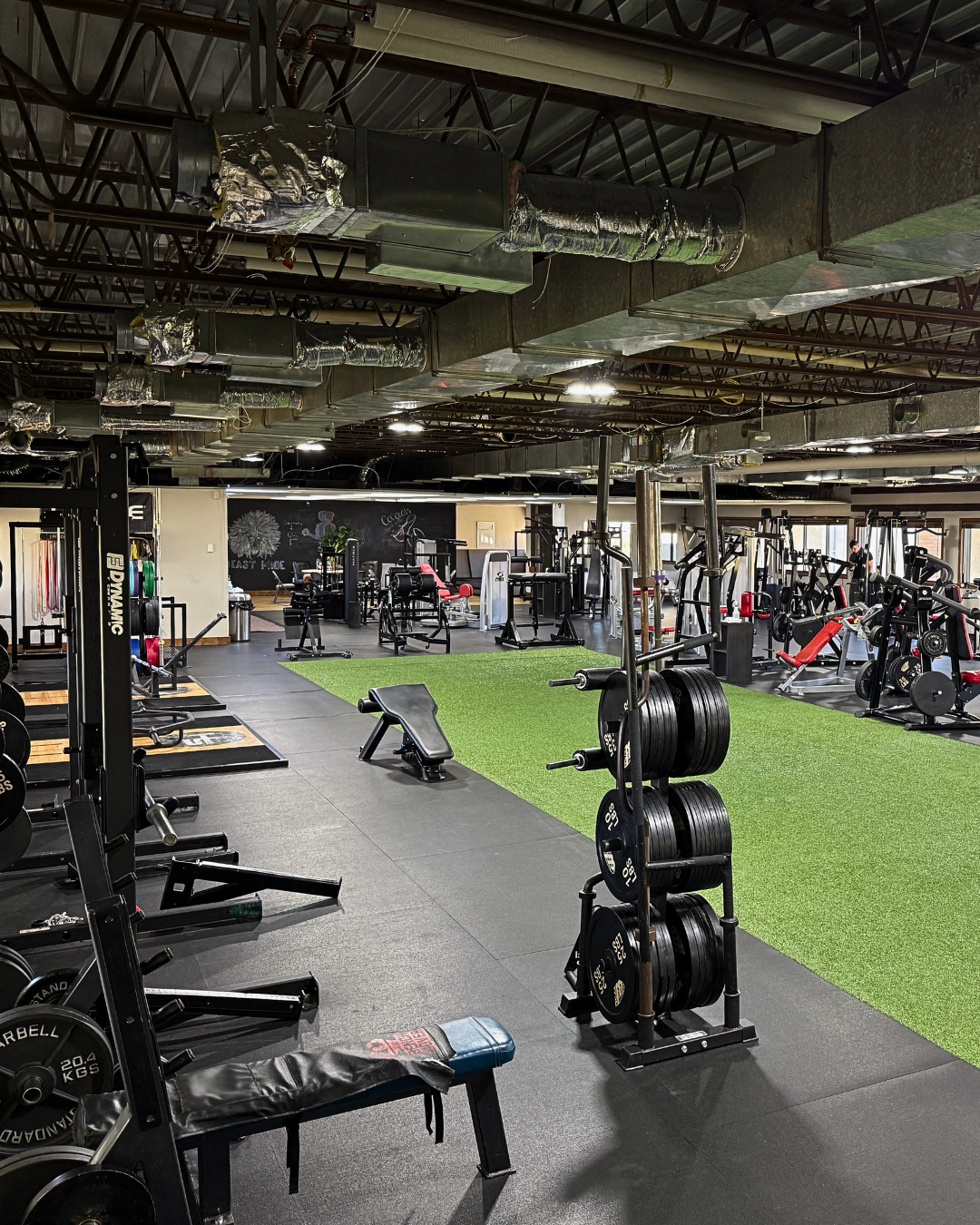 Interior view of a spacious gym with various workout equipment, weight racks, benches, and exercise machines, illuminated by overhead lights and large windows.