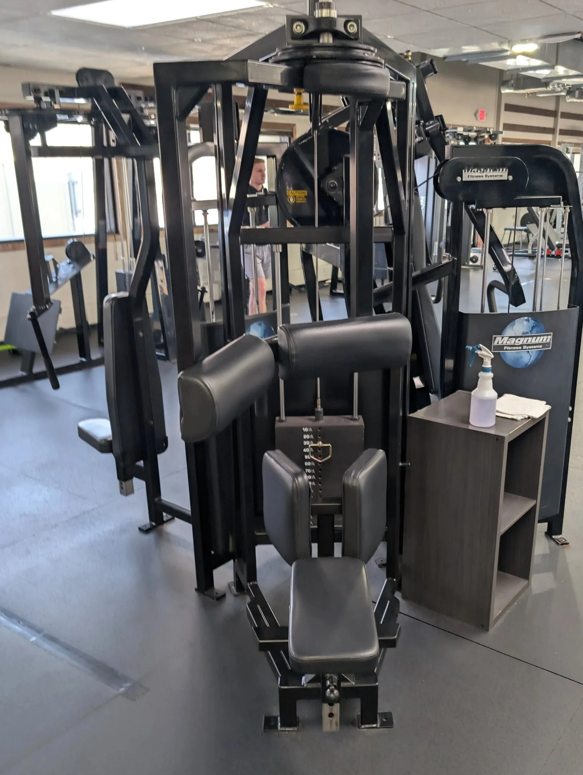 Gym equipment with a black workout machine and a black side table holding cleaning spray and paper towels.