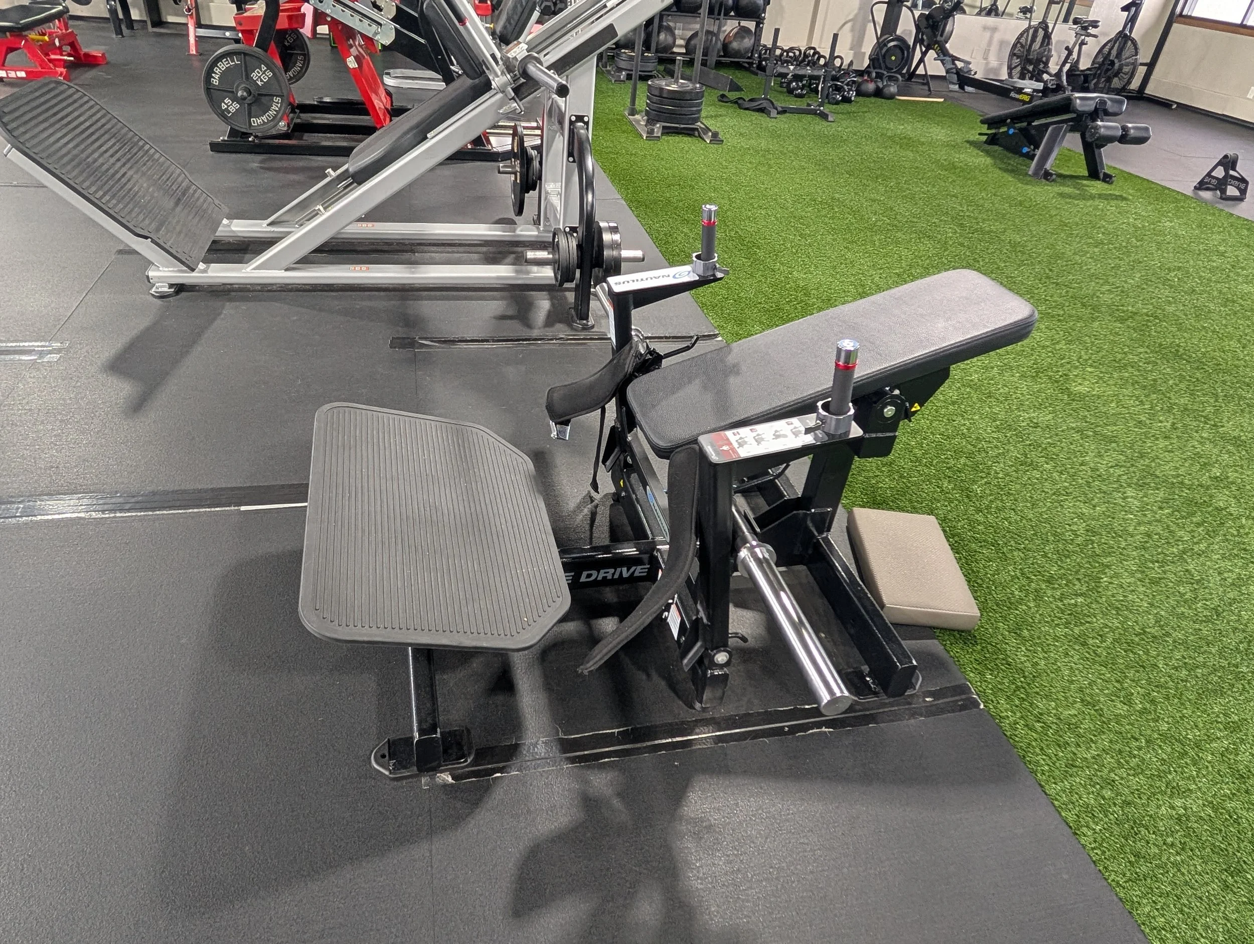 A seated leg curl machine in a gym, with a padded seat and ankle rollers, positioned on black rubber flooring next to artificial turf, surrounded by various gym equipment including weights, benches, and bicycles.