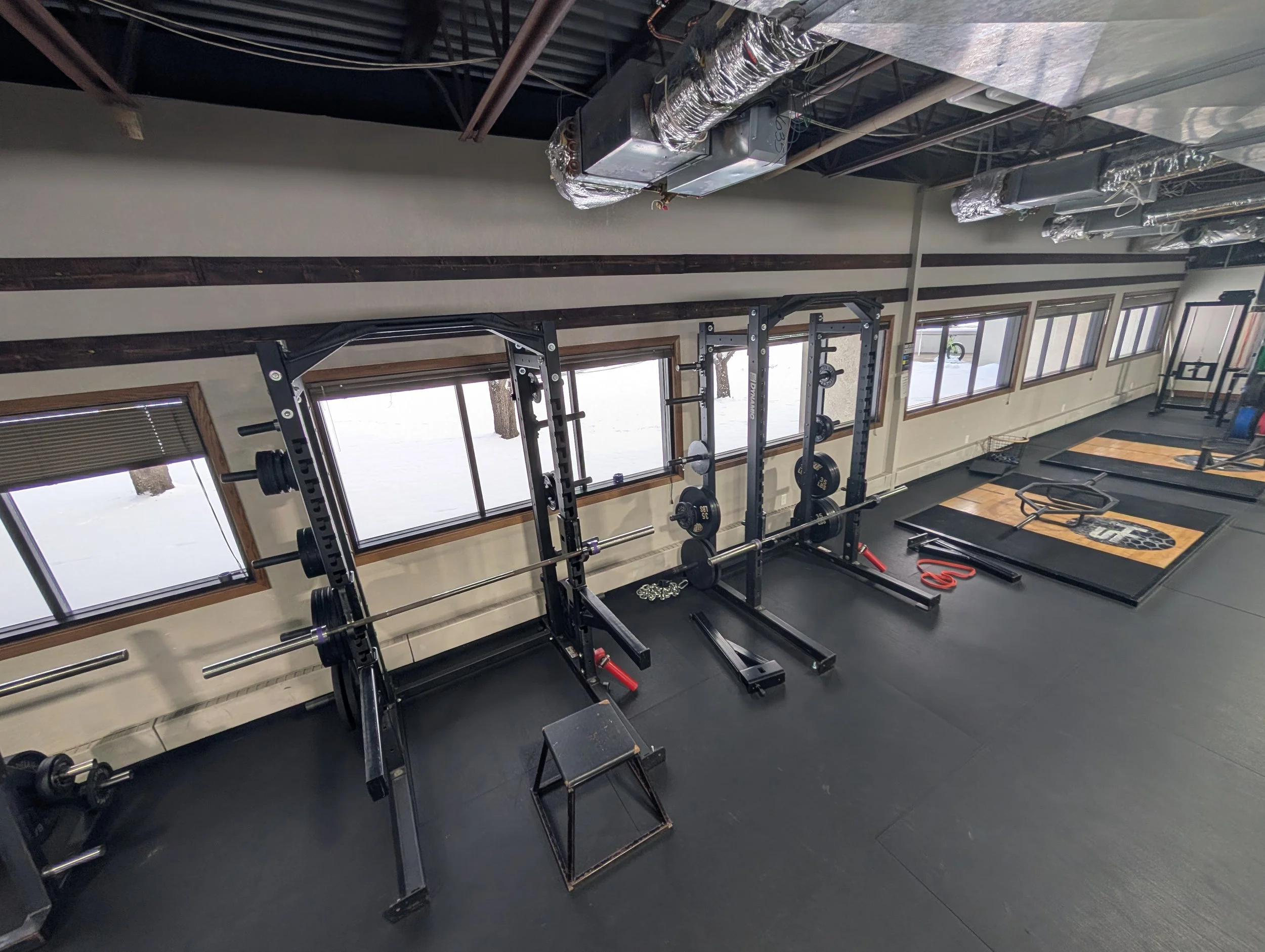 Empty gym with black rubber flooring, weightlifting stations, weight plates, and barbells near windows showing a snowy outdoor scene.