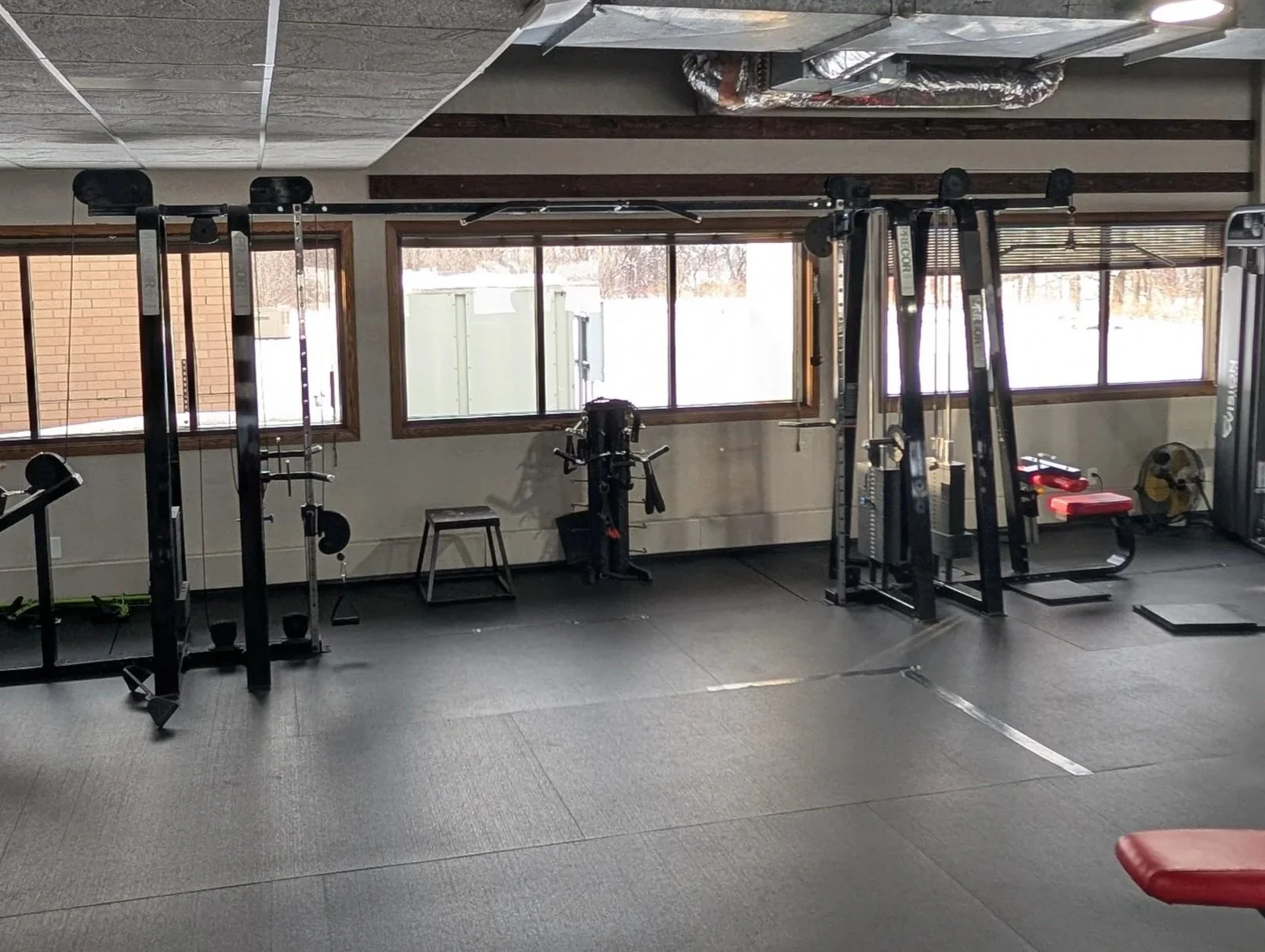 Empty gym with various workout equipment, large windows letting in natural light, and rubber flooring.