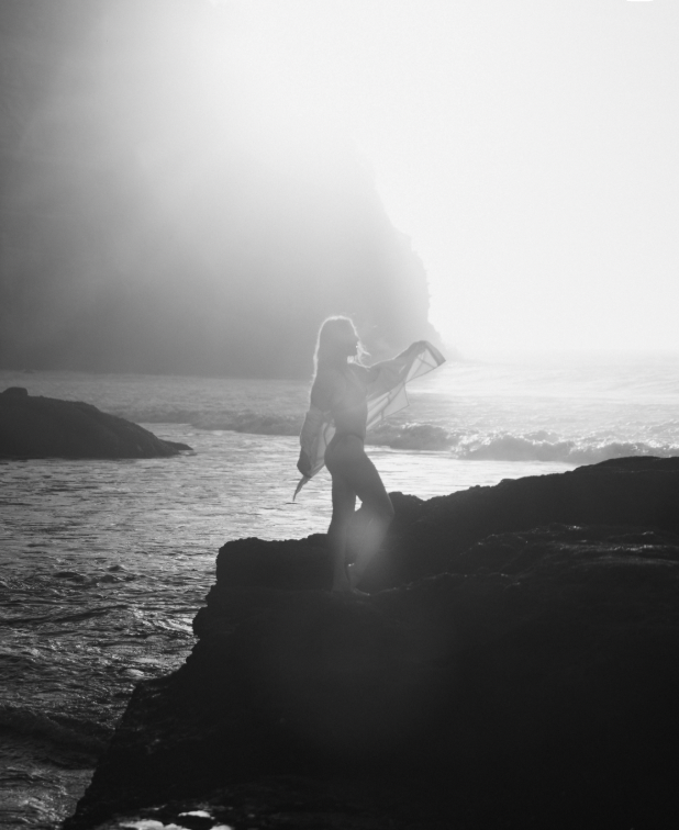 A woman standing on a rocky shoreline, holding a kite surfboard, with the ocean and cliffs in the background during sunset.