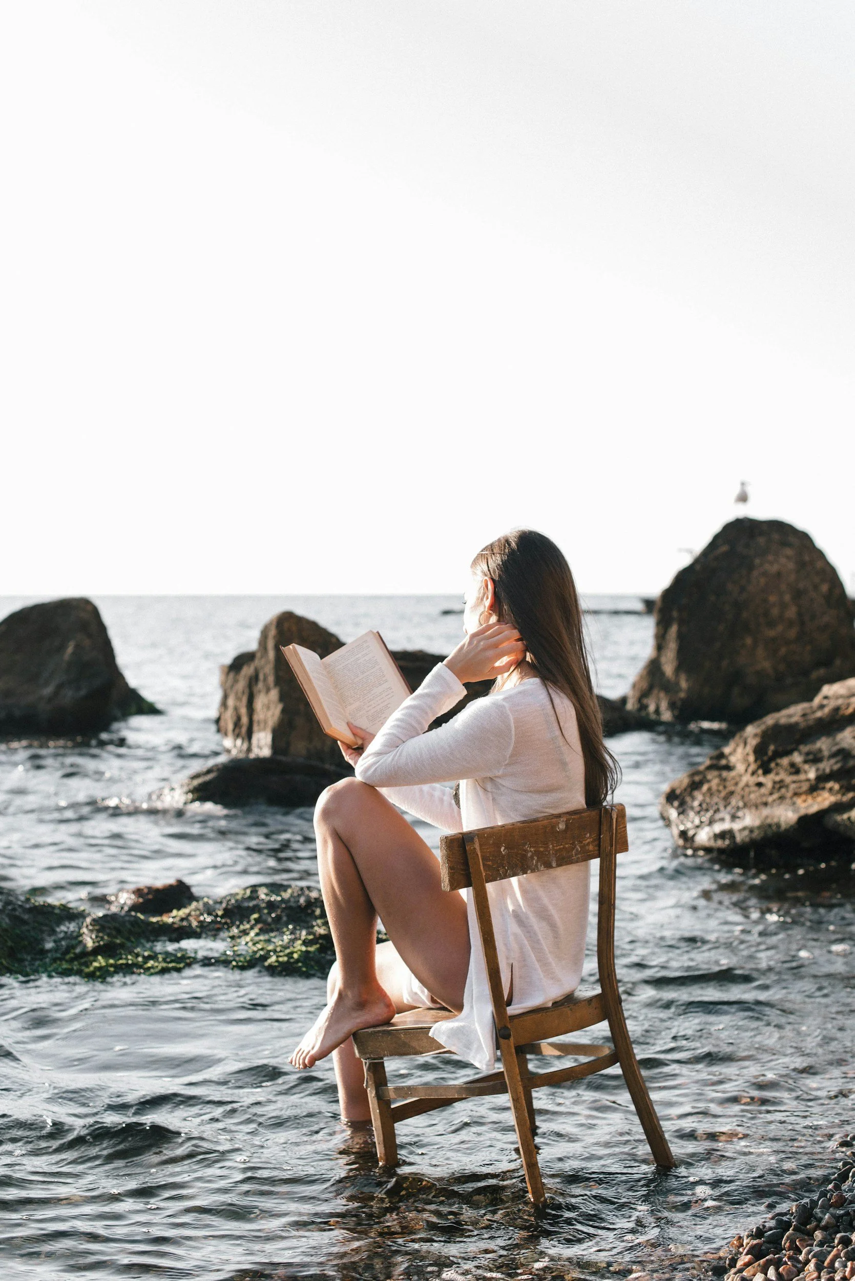 A woman sitting on a wooden chair in the water near rocks, reading a book on a sunny day by the ocean.