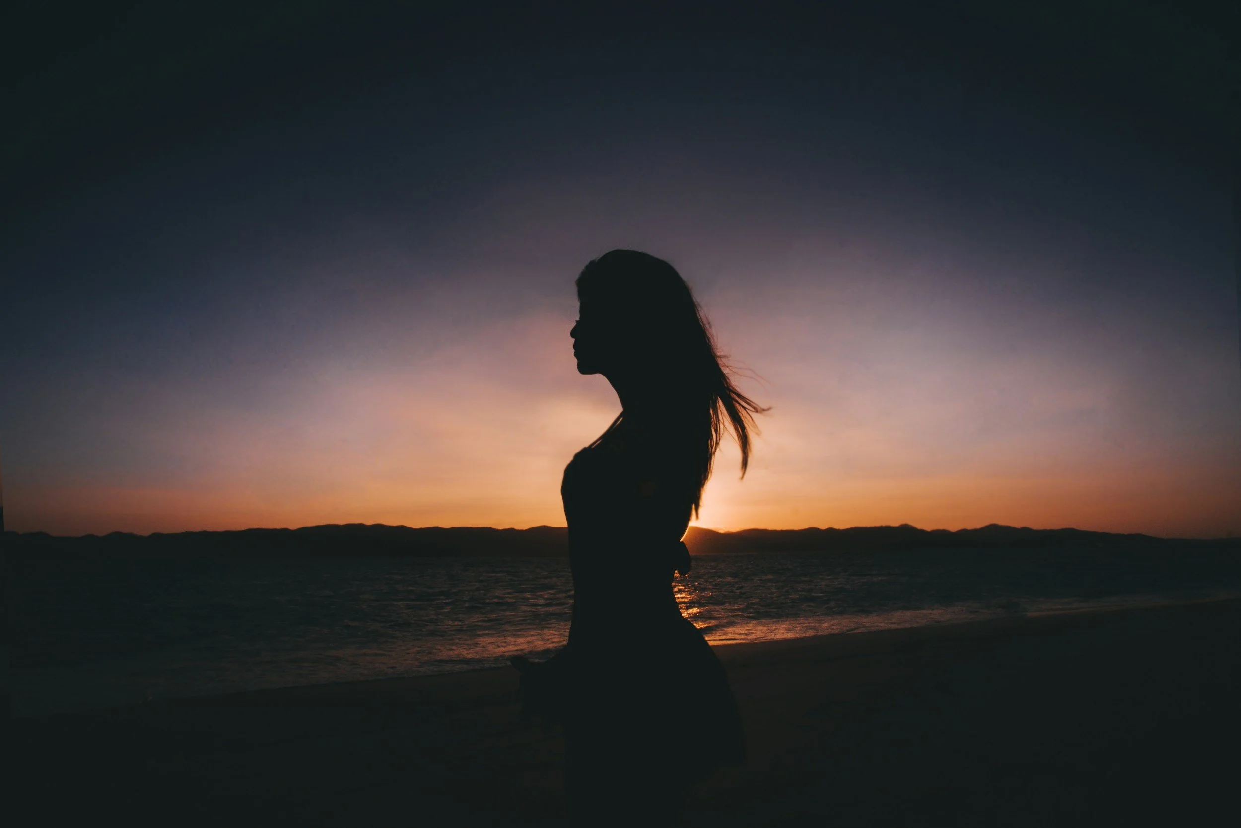 Silhouette of a woman on the beach at sunset, facing left, with the ocean and colorful sky in the background.